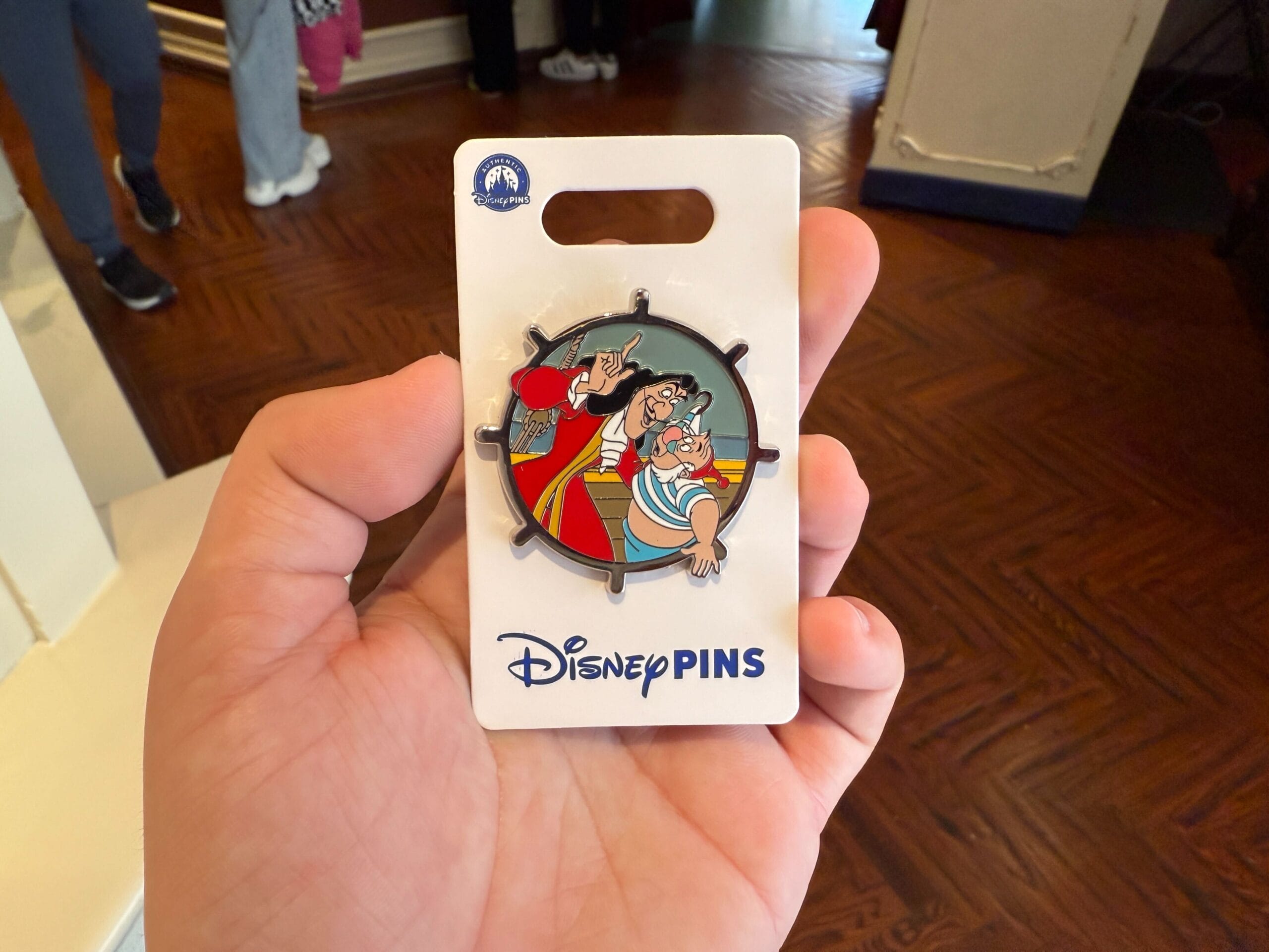 A hand holds a Disney pin featuring two animated characters, one in red with a hook hand and the other in blue and white stripes. The card labeled "Disney Villains Pins" hints at its exclusive availability, making it a cherished find at Disneyland Resort.