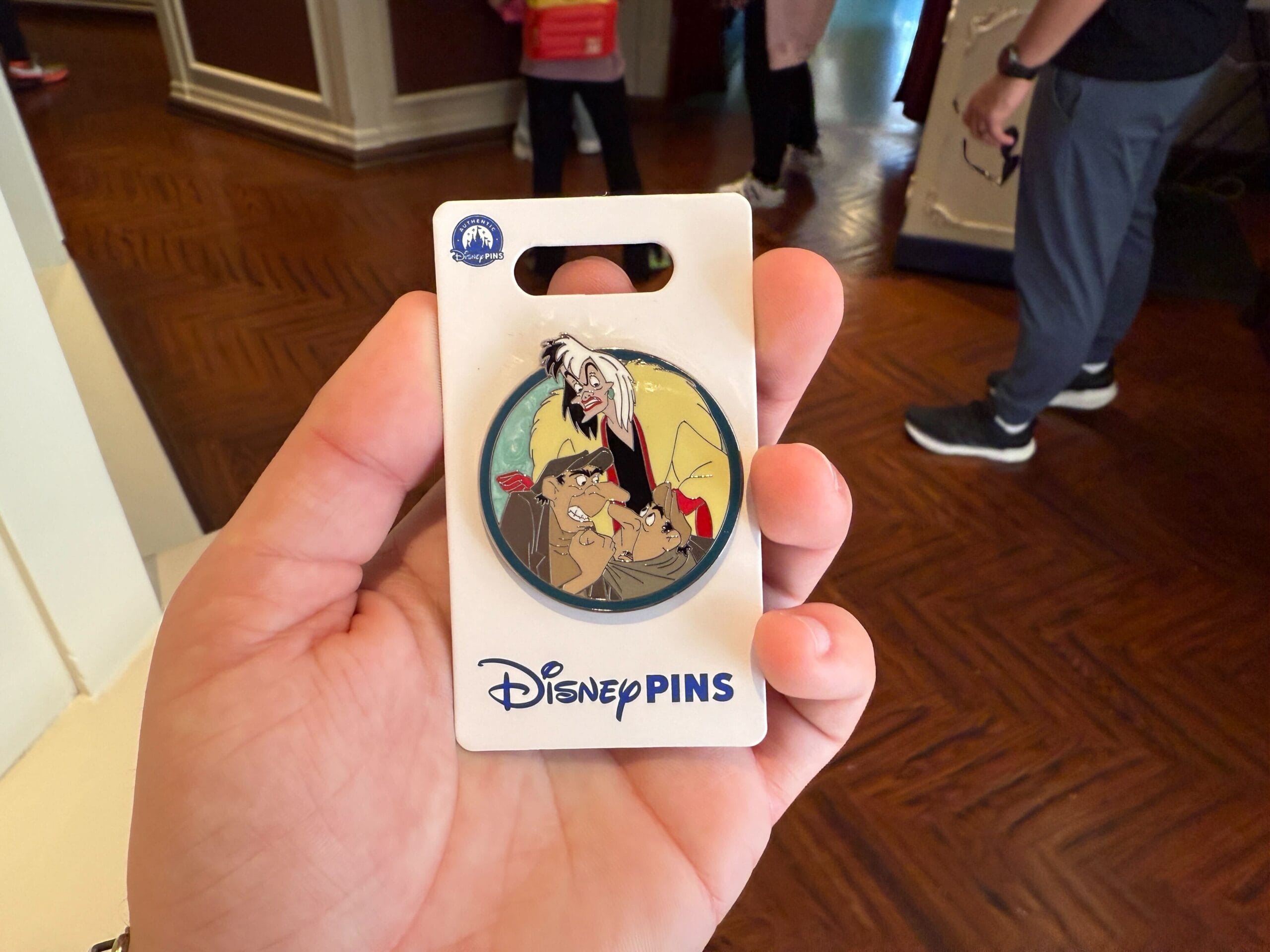 A hand holds a Disney Villains Pin featuring a cartoon character with two spotted animals, attached to a labeled card. Amid the bustling background of the Disneyland Resort, people and wooden flooring enhance the lively atmosphere as new pins are showcased.