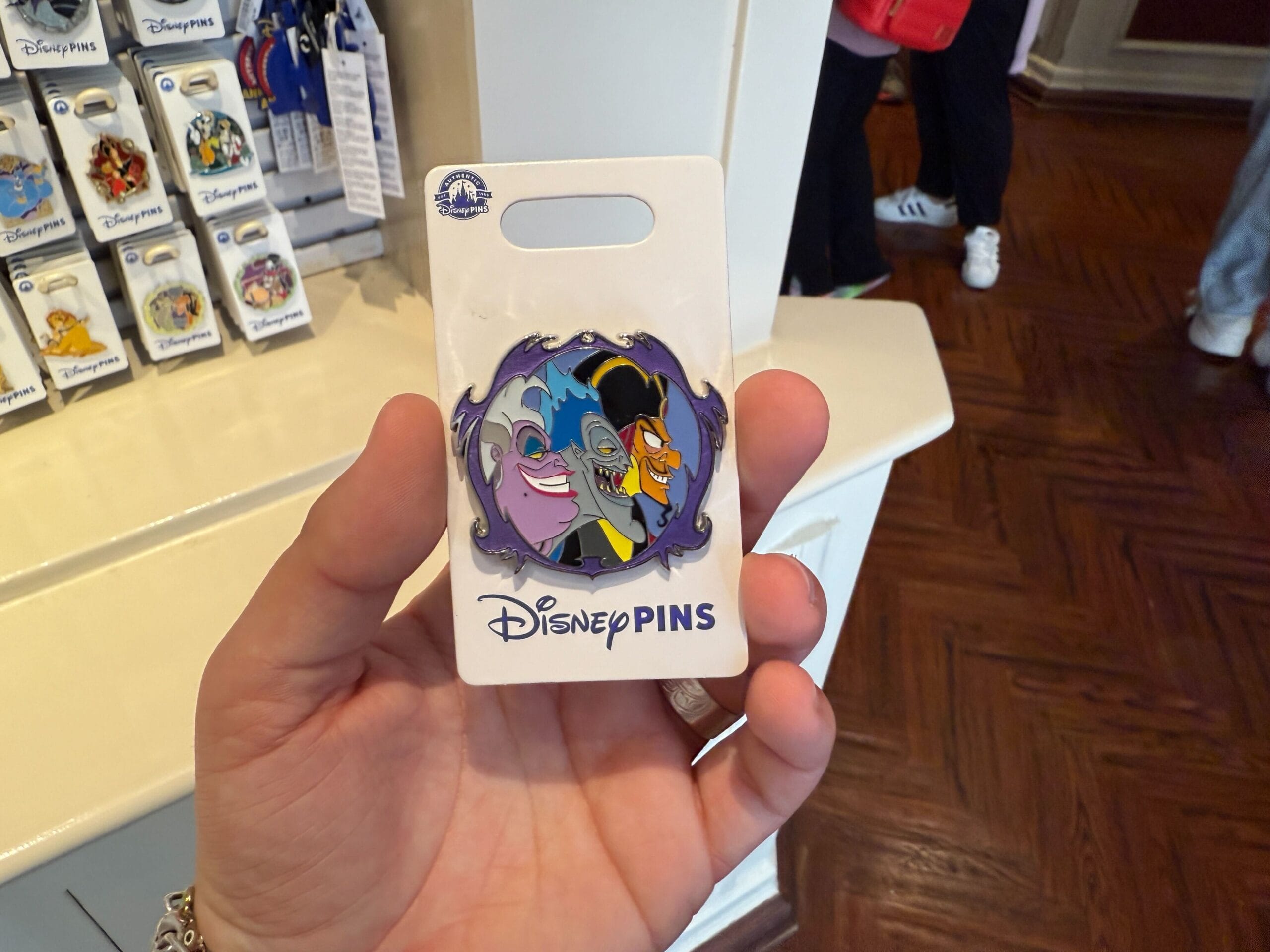A hand holds a Disney Villains Pin featuring cartoon characters with an evil grin, showcased on packaging that reads "Disney PINS." In the background, you can glimpse a retail display and wooden floor, reminiscent of the enchanting atmosphere at Disneyland Resort.