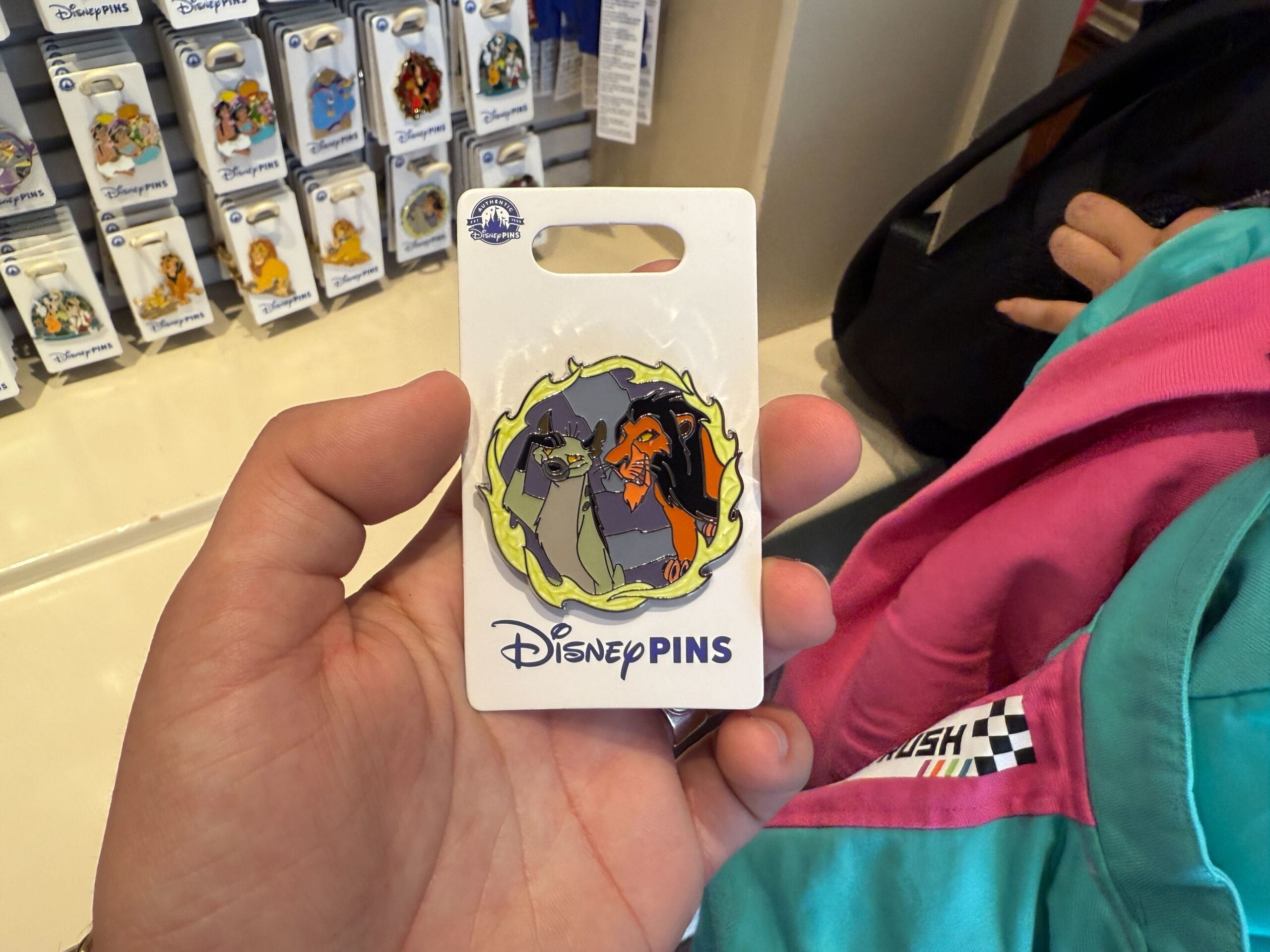 A hand holds a Disney Villains Pin featuring Scar and a hyena from "The Lion King," attached to a white card labeled "Disney Pins." Among the new pins at Disneyland Resort, various other pins are displayed in the background.
