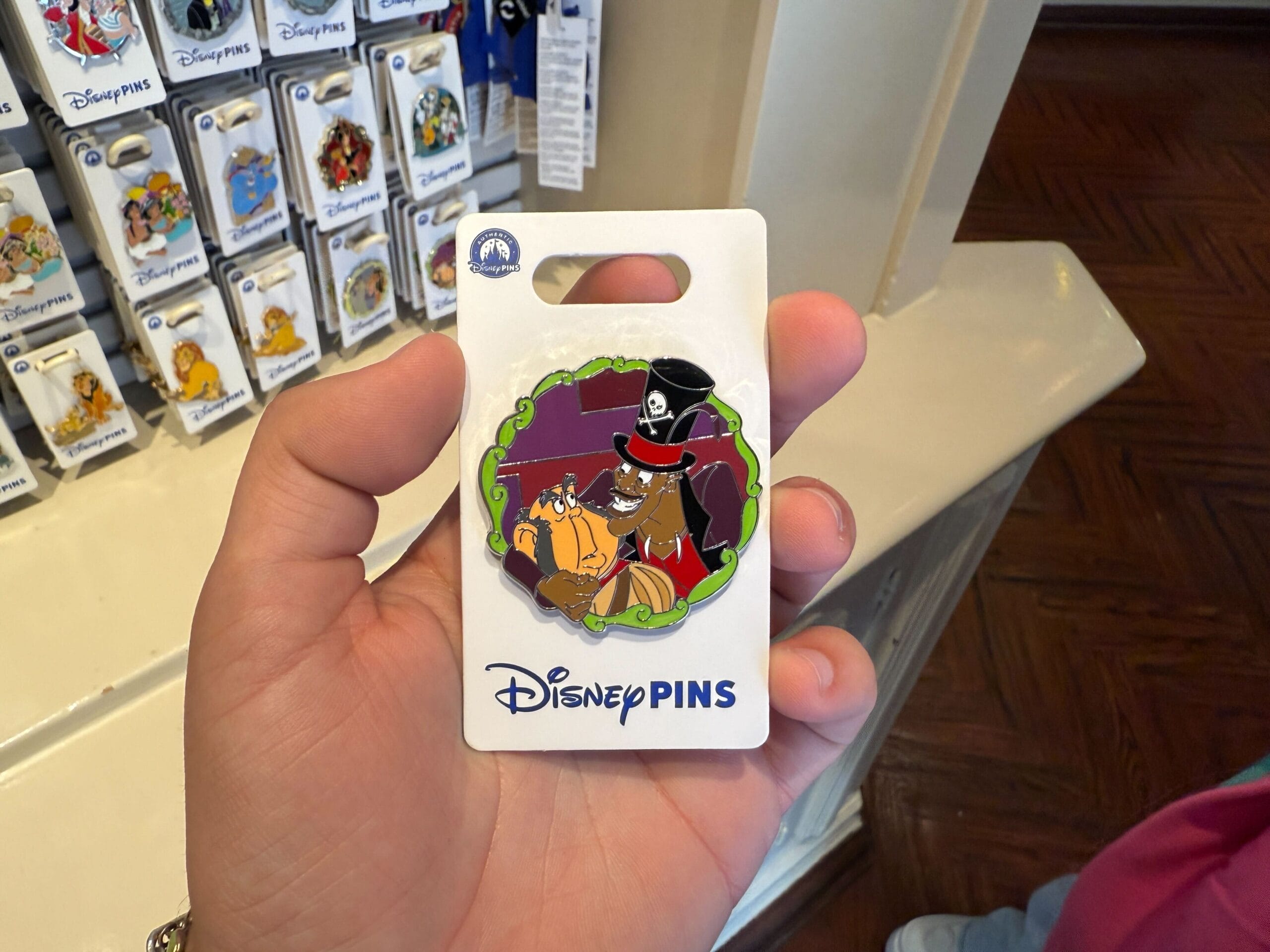 A hand holds a Disney pin featuring an animated pirate dog and a scared cat inside a store display area at Disneyland Resort. Other new pins shimmer in the background, adding to the vibrant collection.