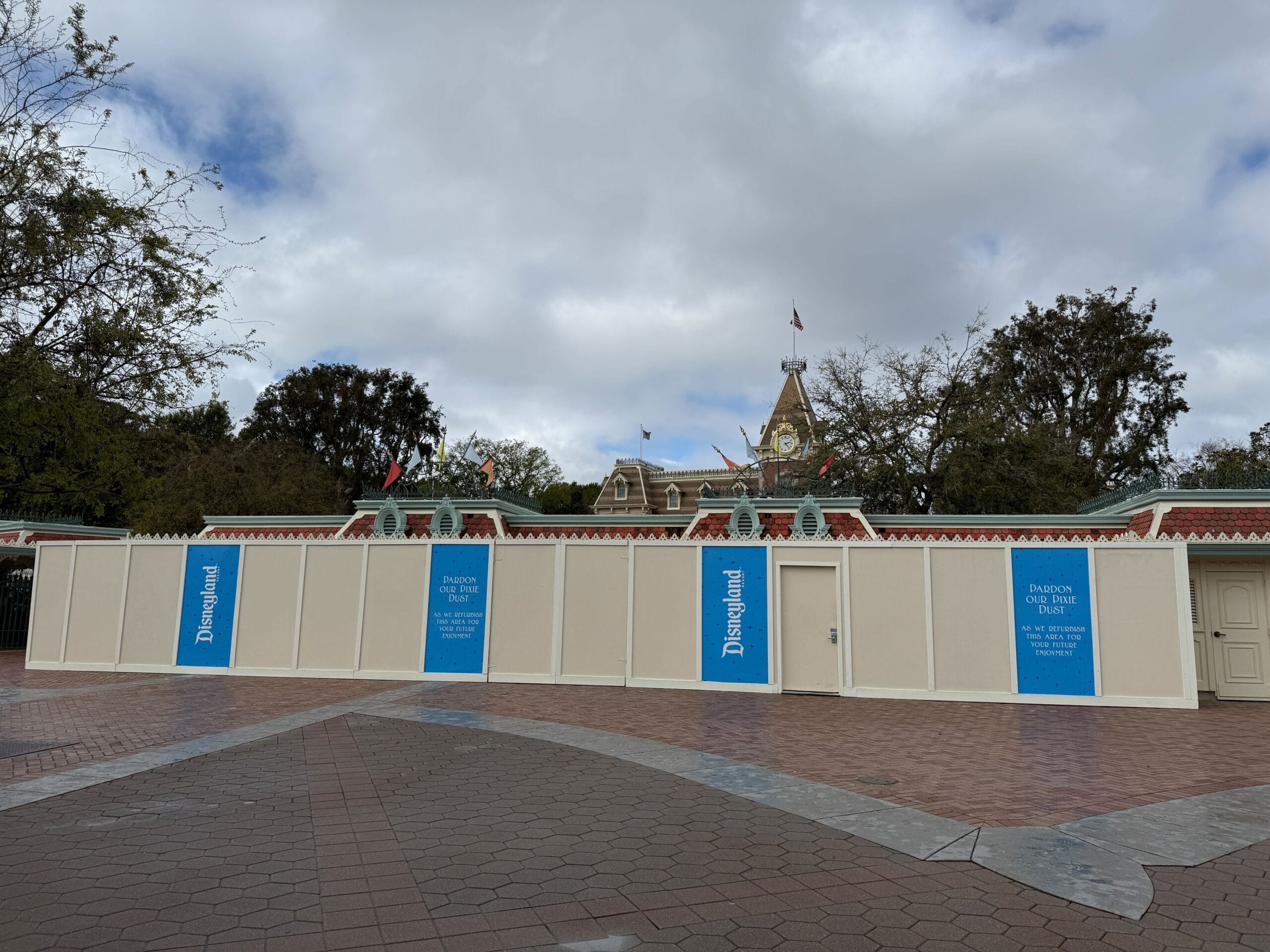 Construction walls with Disneyland logos and blue signs are set up in front of a park entrance building with a clock tower. The sky is cloudy, and there are trees in the background.