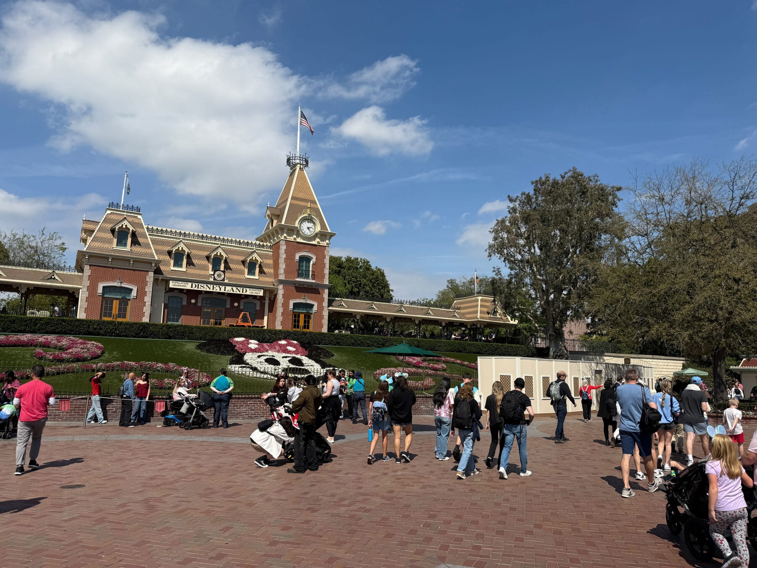 People gather in front of Disneyland entrance, featuring a floral Mickey Mouse design. Blue sky and clouds are visible.