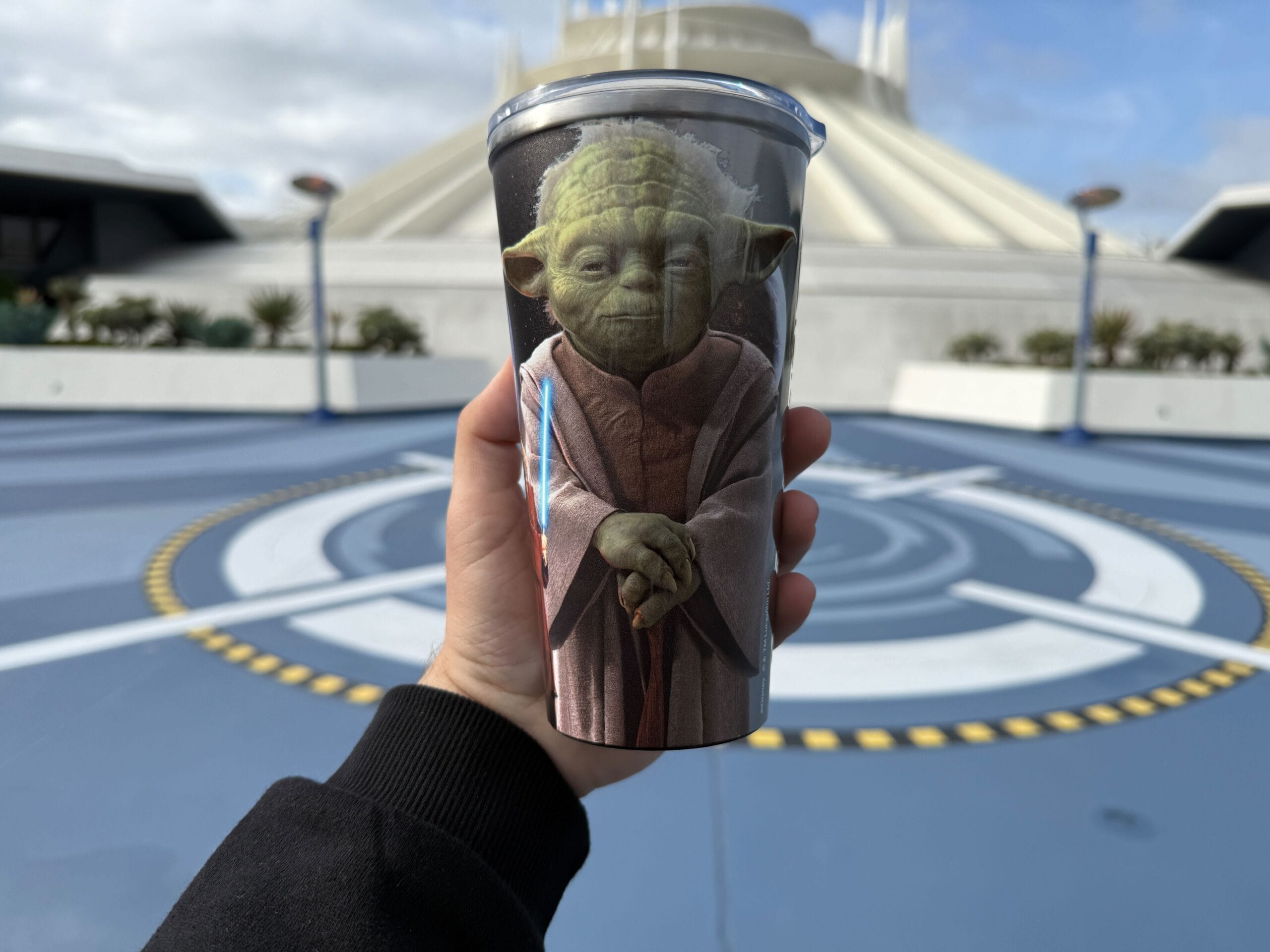 A person holds a cup featuring an image of a green, robed character with large ears in front of a futuristic building under a blue sky.