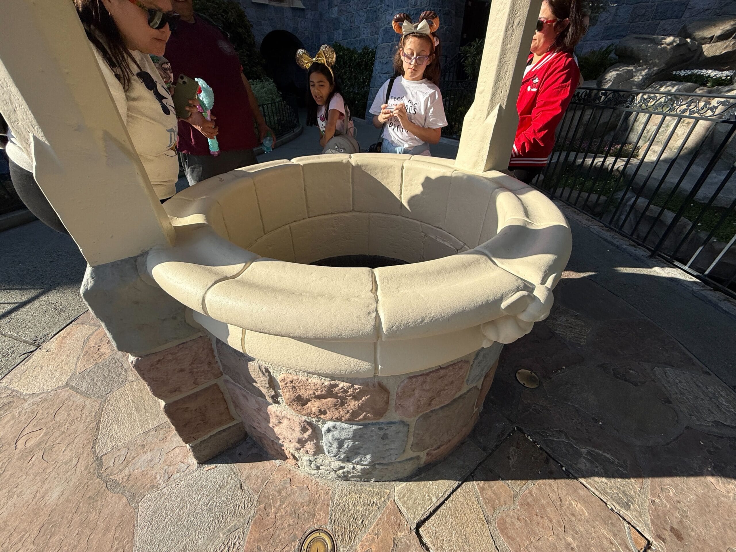 Visitors gather around a stone wishing well at the amusement park, with some holding phones and wearing themed accessories. It's reminiscent of the enchanted experience at Disneyland Resort, where stories like Snow White come to life.