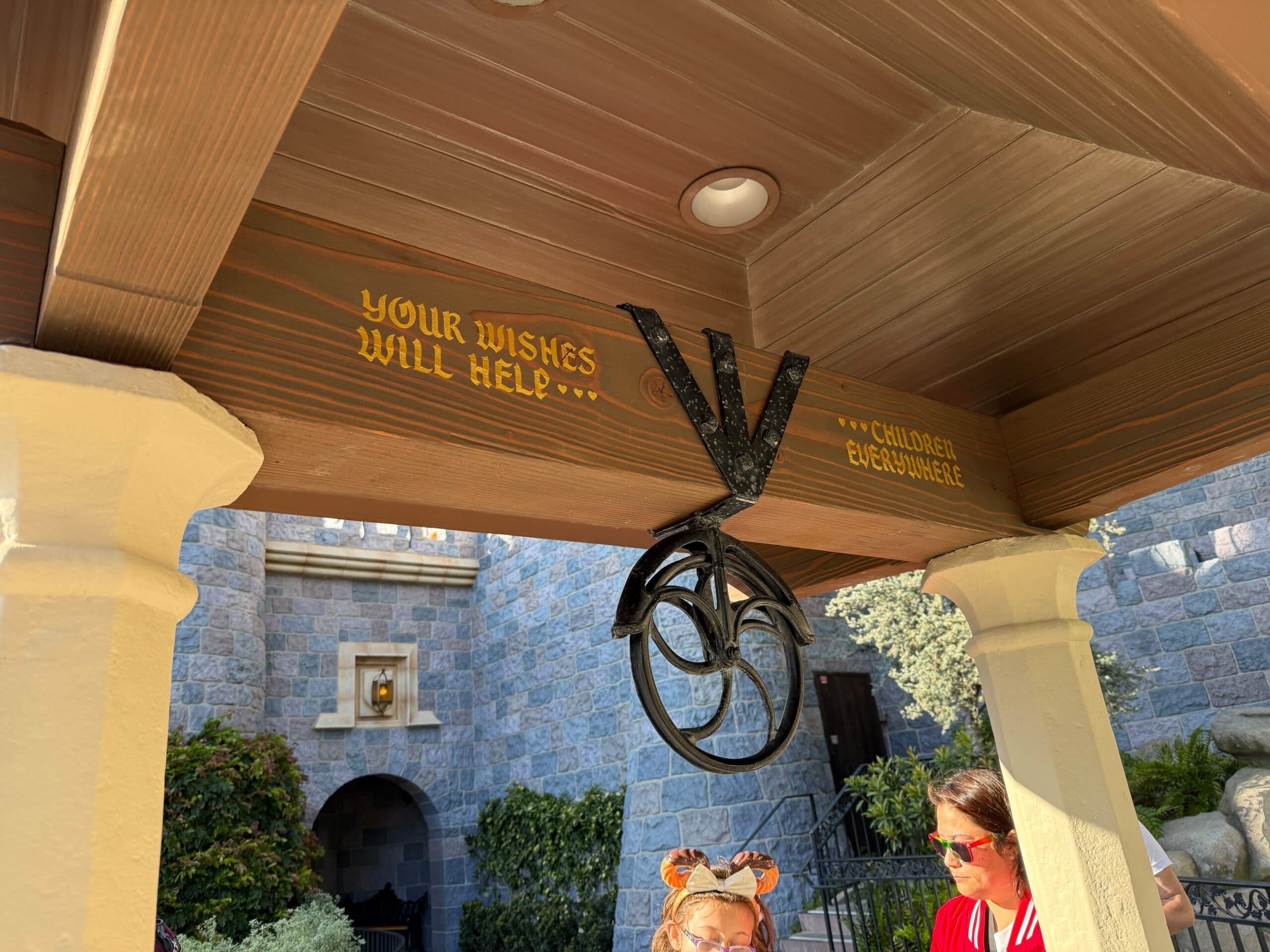 A wooden beam in a castle setting, reminiscent of Snow White's Wishing Well at Disneyland Resort, displays the engraved message "Your wishes will help... children everywhere." Two people are visible below the beam, gazing up hopefully.