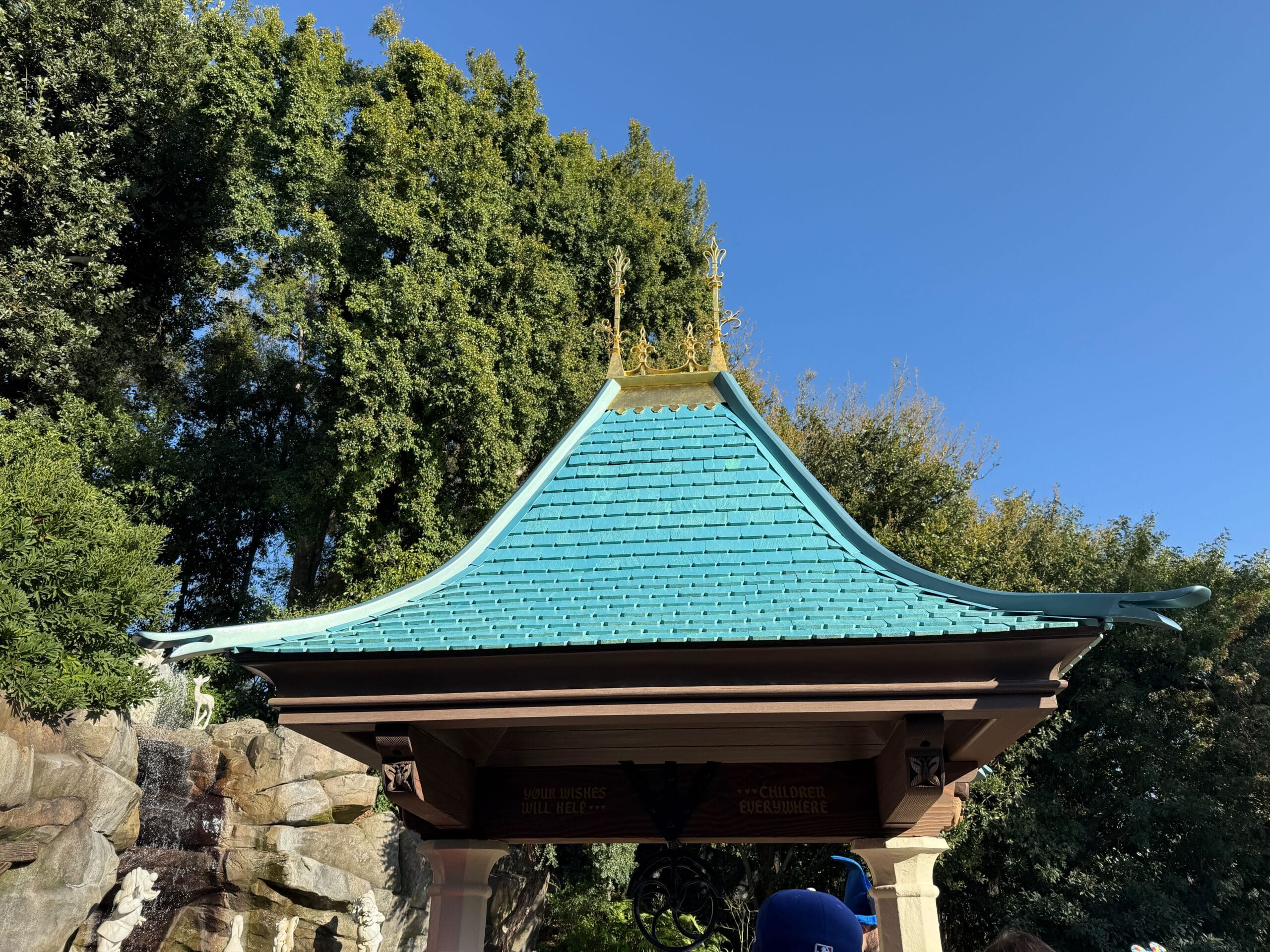 The freshly-repainted roof of the pagoda-style building glistens with teal and ornate golden details against a clear blue sky, reminiscent of a fairytale scene near Snow White's Wishing Well, surrounded by verdant trees.