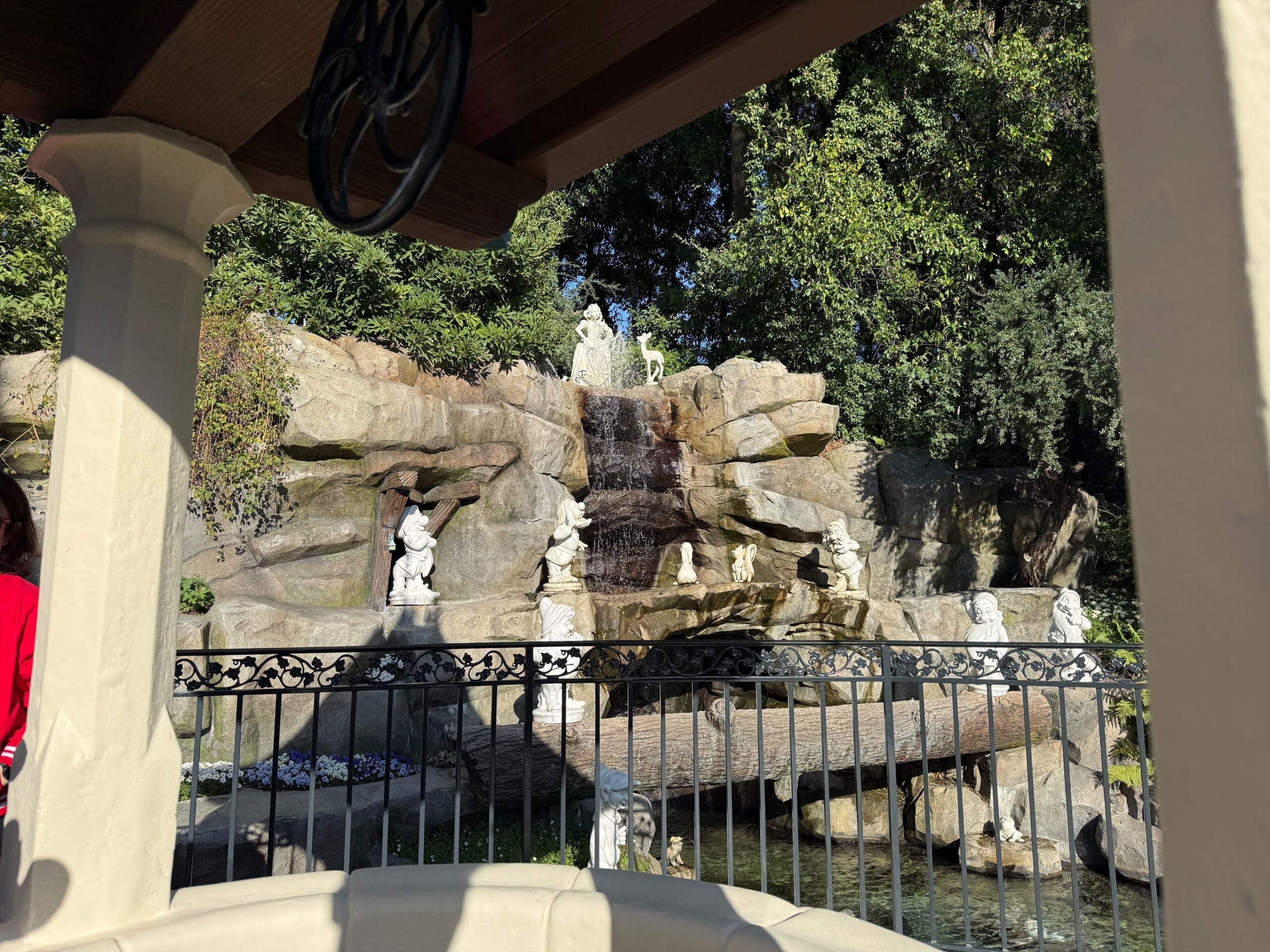 A stone waterfall feature with various statues of animals and figures is behind an ornate fence in a wooded garden setting, reminiscent of Snow White's Wishing Well at Disneyland Resort.