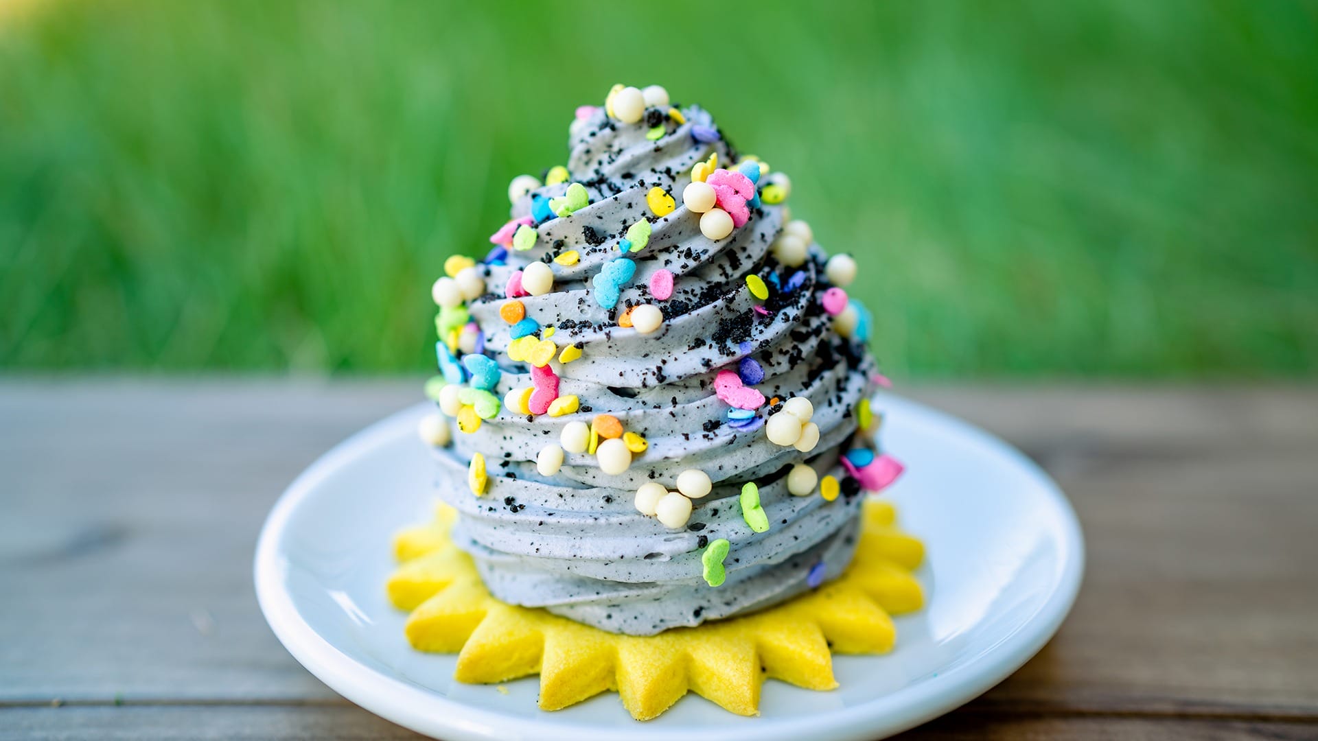 A swirl of gray ice cream topped with colorful sprinkles sits on a white plate with a yellow, flower-shaped cookie, against a grassy background.