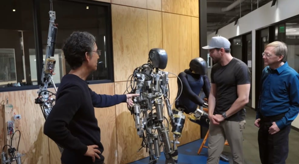 Three people observe a partially assembled humanoid robot in a room with wooden walls, possibly dreaming of Disneyland Paris's visionary tech. In the background, another Stuntronic awaits its moment to perform.