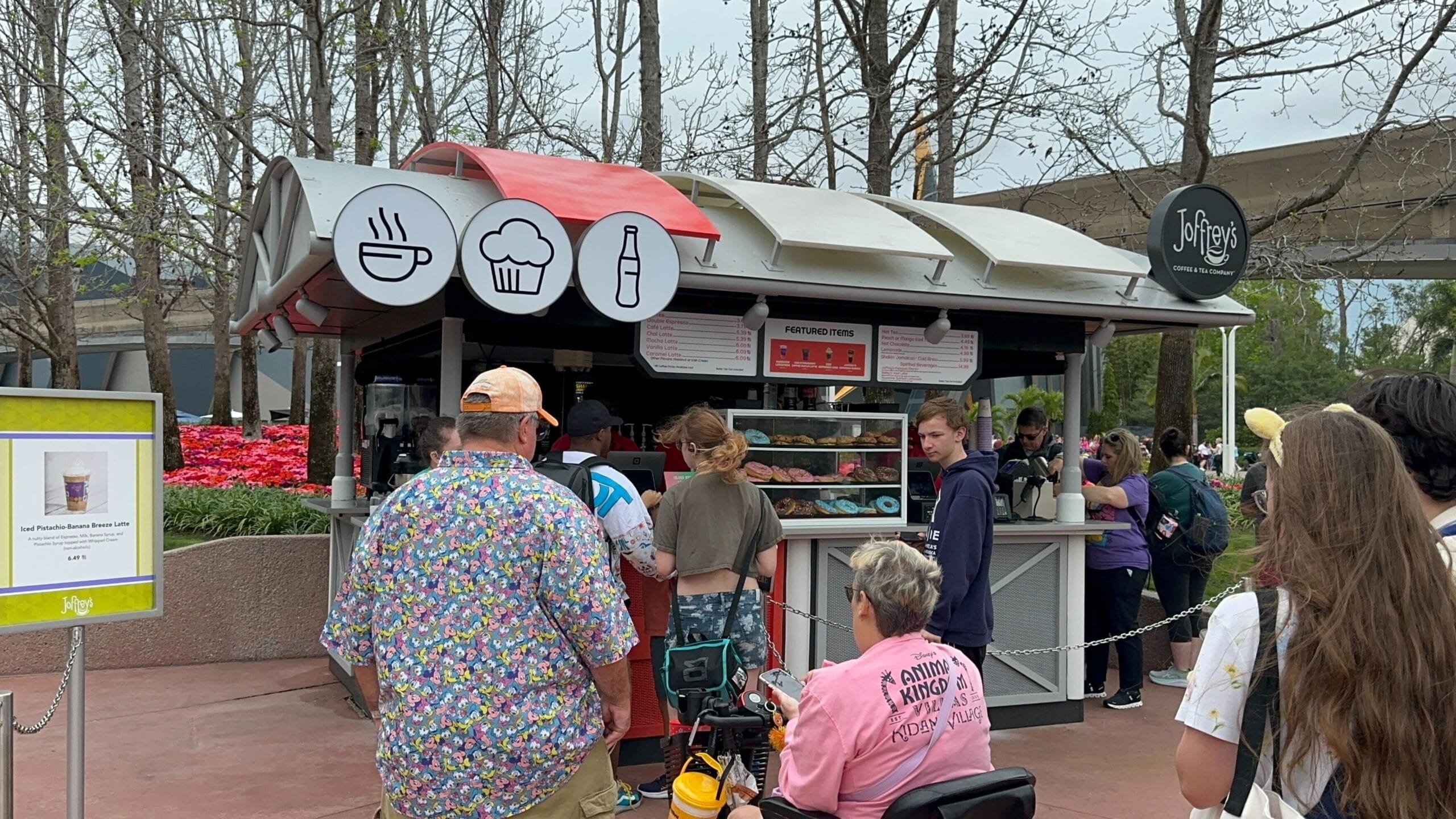 People lined up at Joffrey's food kiosk, with signs depicting a burger, cupcake, and drink. A menu board is visible, while some individuals linger near flowers and trees in the background.
