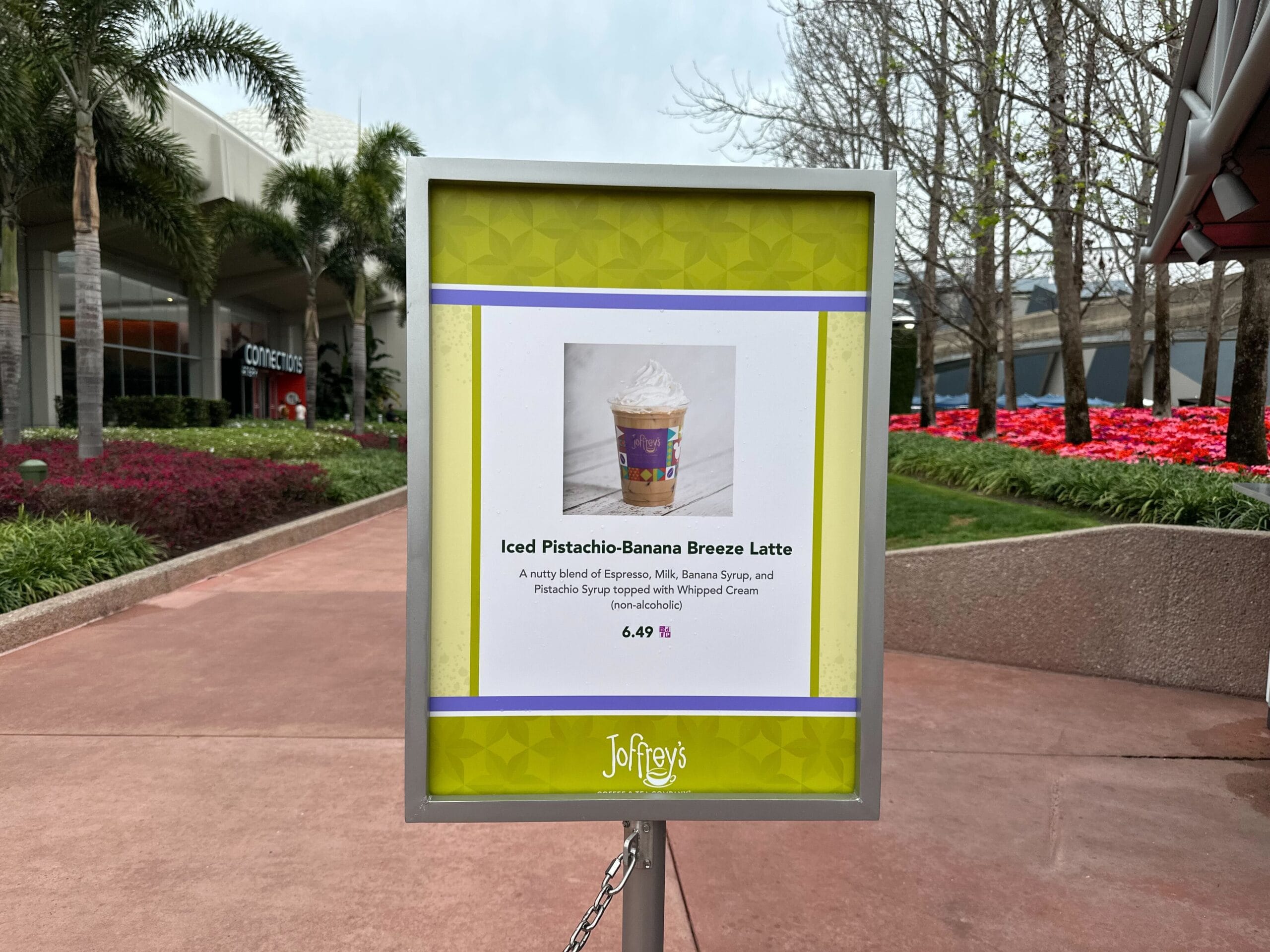 Discover the Joffrey's iced pistachio-banana breeze latte for just $6.49. This refreshing delight comes with an enticing photo and description, set against a backdrop of trees, red flowers, and a concrete pathway.
