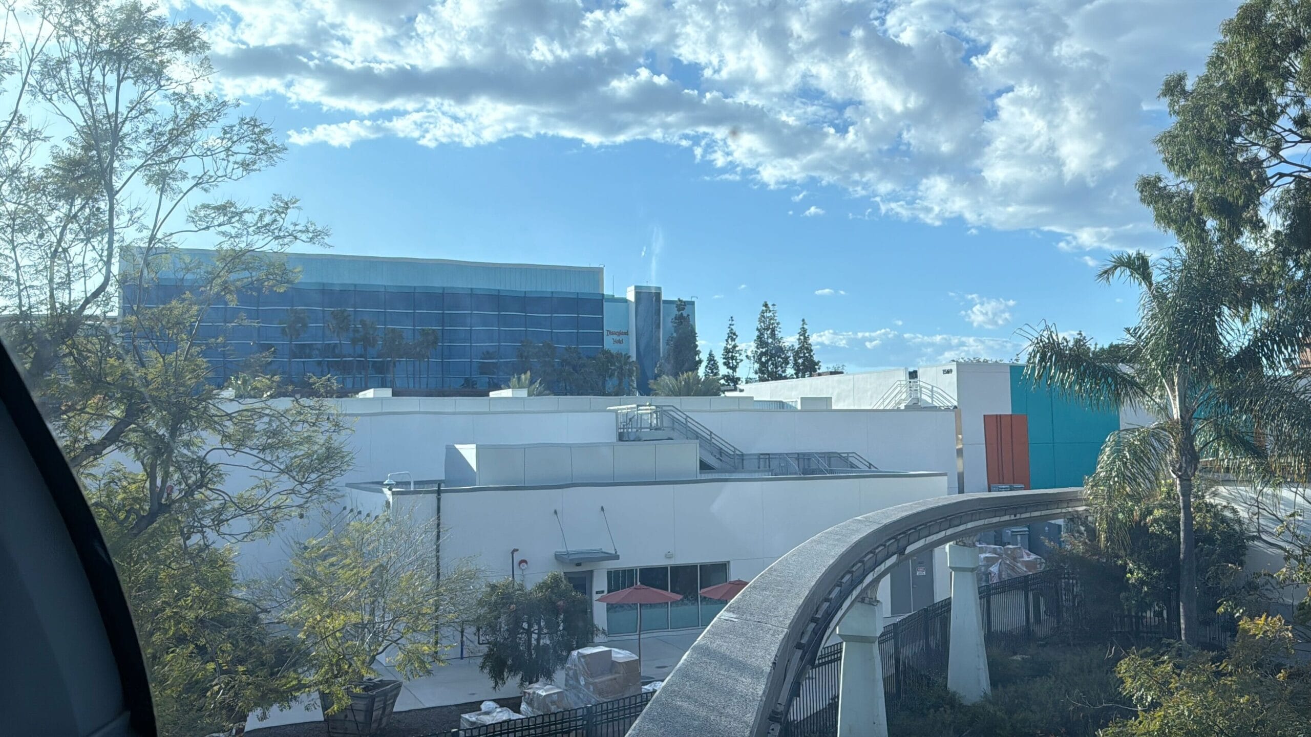 The Disneyland Monorail track with support columns gracefully passes through a landscaped area filled with trees, adjacent to modern buildings under a clear, partly cloudy sky. Recently reopened, it offers a seamless journey through scenic and urban landscapes alike.