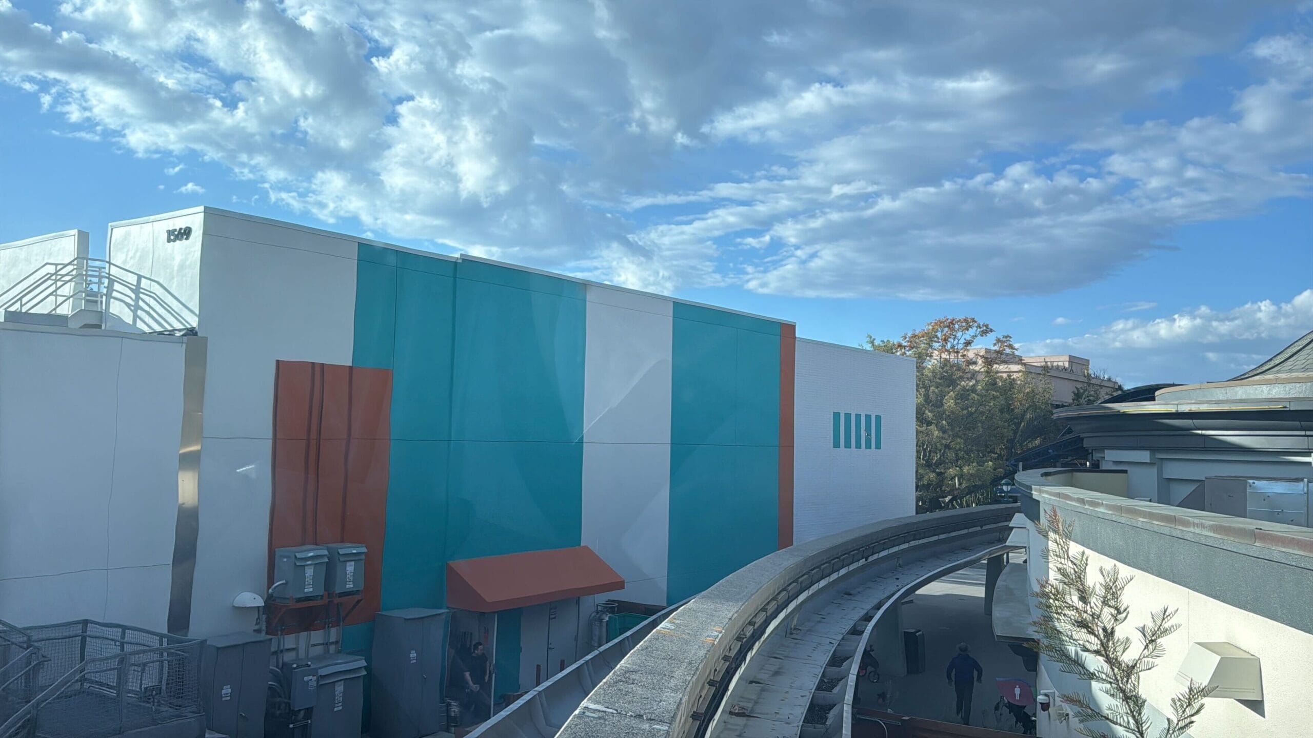 A building with teal, white, and orange panels stands under a cloudy sky, while the recently reopened Disneyland Monorail track curves gracefully in the foreground.
