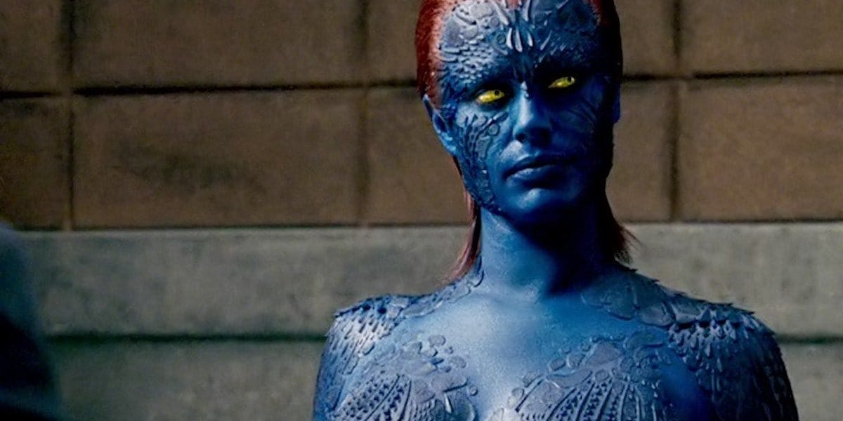 Against a stone backdrop, a figure resembling Mystique from the Marvel Cinematic Universe stands with blue, textured skin and yellow eyes, accented by short red hair.