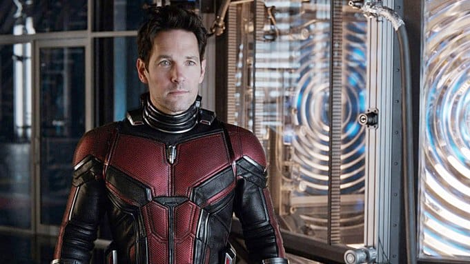 In a futuristic lab filled with metal and glass equipment, Ant-Man stands confidently in his red and black superhero suit, marking his anticipated return to the thrilling adventures of the Marvel Cinematic Universe.