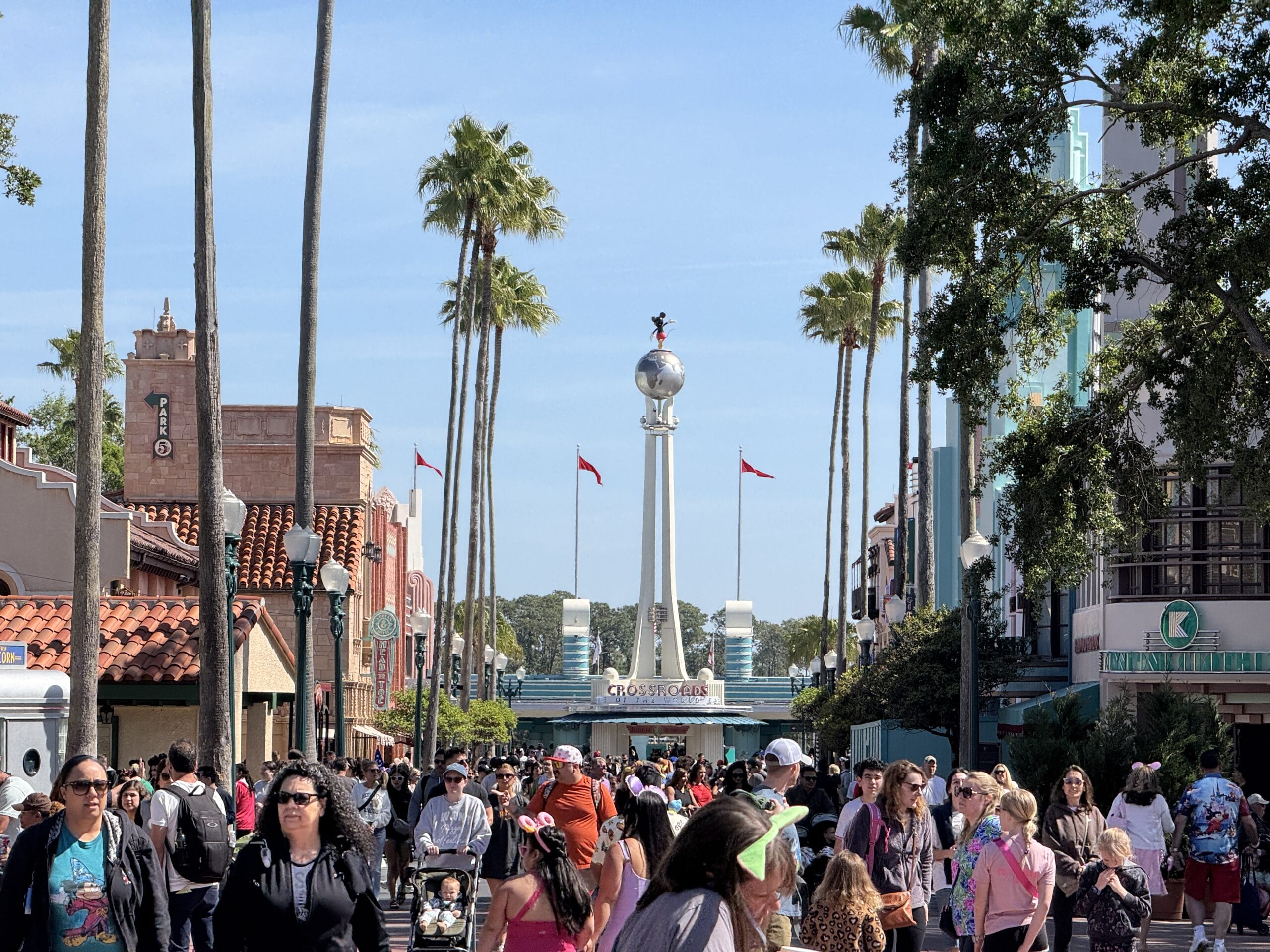 Amid a bustling scene at Disney's Hollywood Studios, with towering palm trees and an ornate tower in the backdrop, vacationers stroll along the sunlit pathways, capturing memories for their photo report.