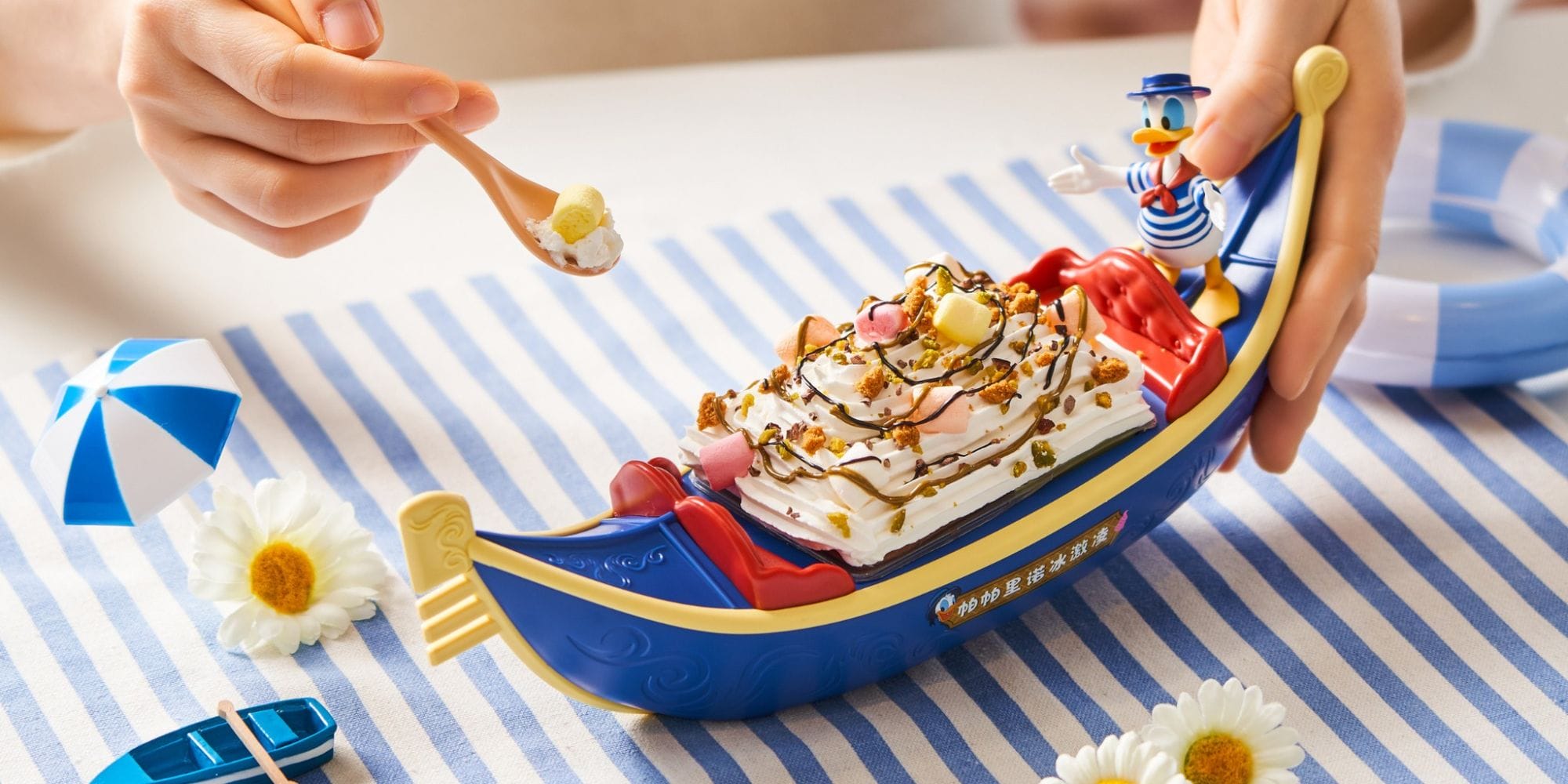 A person holding a spoonful of dessert from a Donald Gondola Cup, shaped like a gondola with a beloved character, sits on a striped cloth adorned with daisies and a mini umbrella. Enjoy this whimsical treat at Il Paperino Ice Cream Parlor in Shanghai Disneyland.