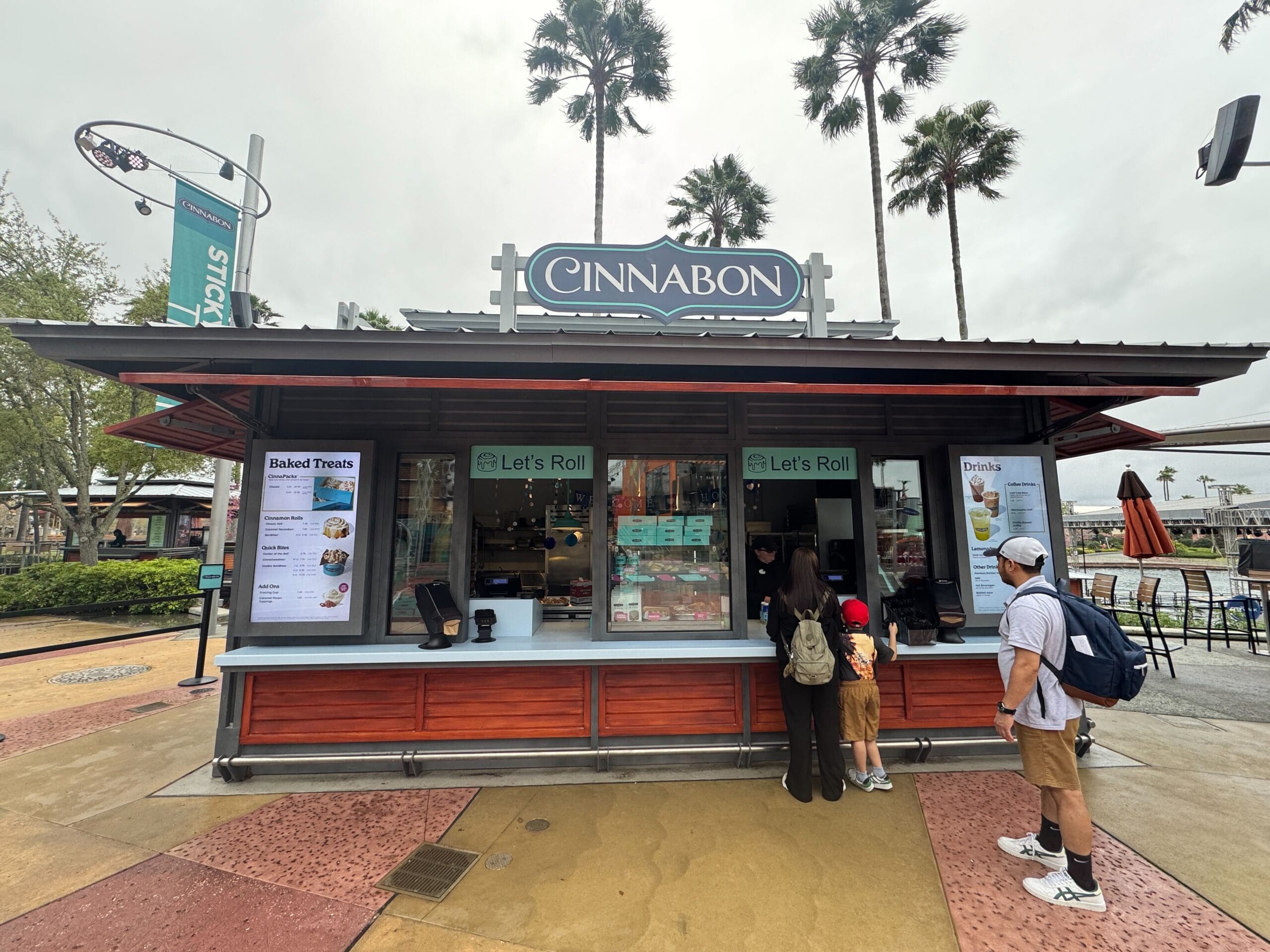 People standing at the counter of a Cinnabon kiosk with "Let's Roll" signs catch a sweet break at Universal Orlando's CityWalk. Palm trees sway subtly in the background under an overcast sky, adding to the relaxed vibe of this iconic spot.