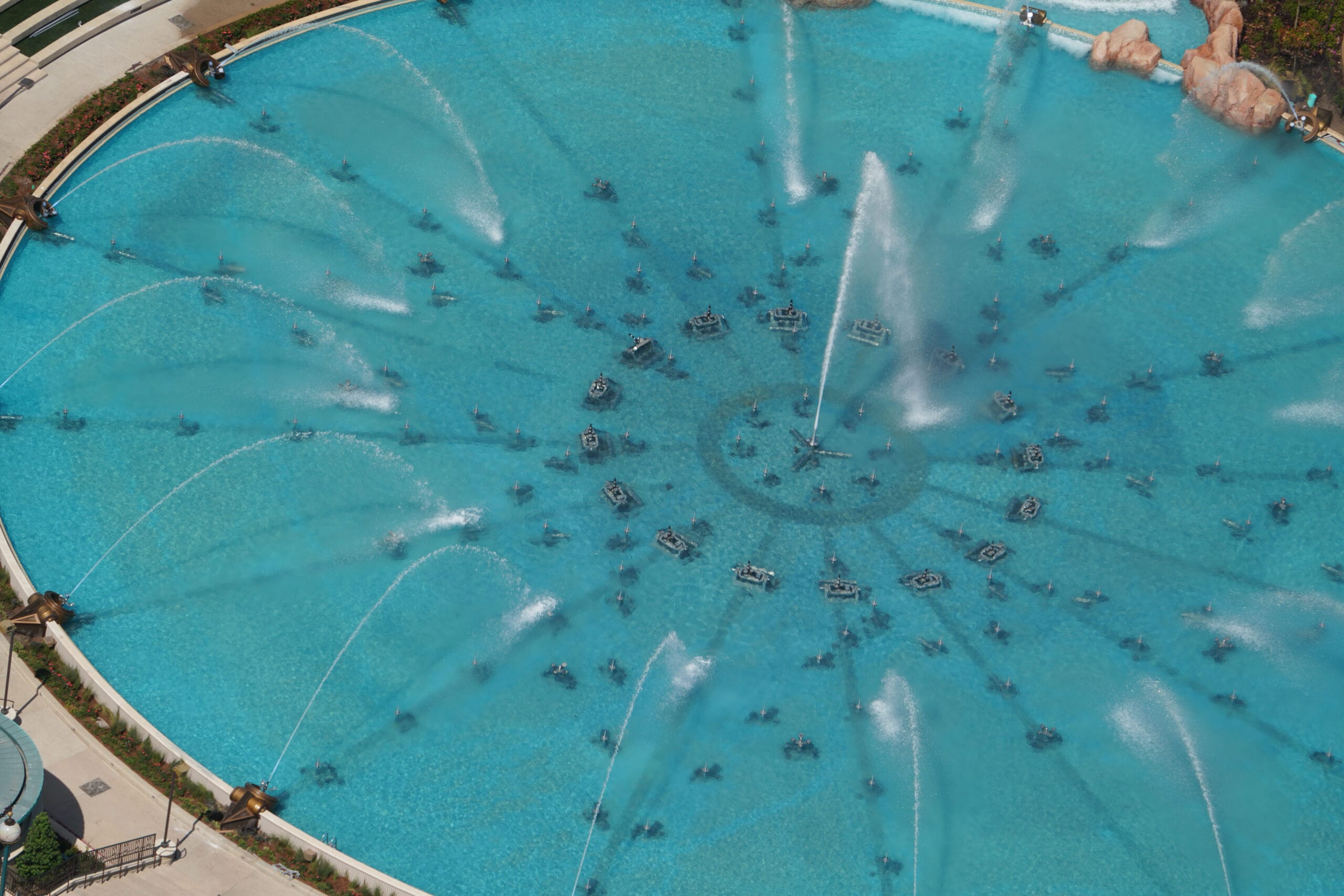Aerial view of a large circular fountain undergoing testing, with multiple water jets creating symmetrical patterns in a clear blue pool. Concrete pathways encircle the fountain.