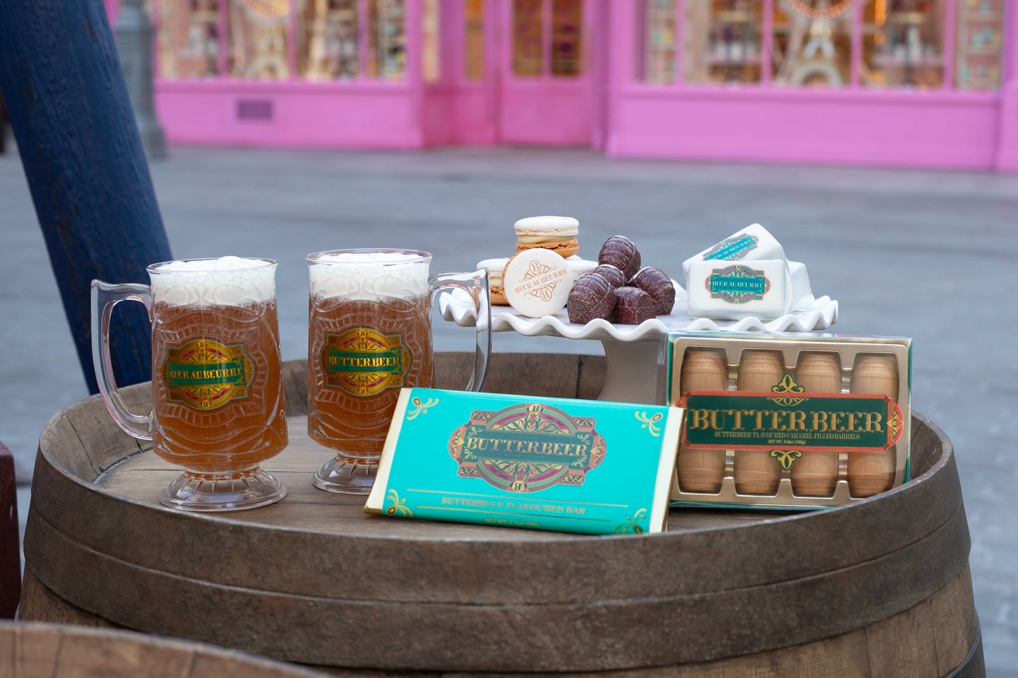 Two mugs of frothy butterbeer and packages of sweets on a wooden barrel, with a pink shopfront in the background.