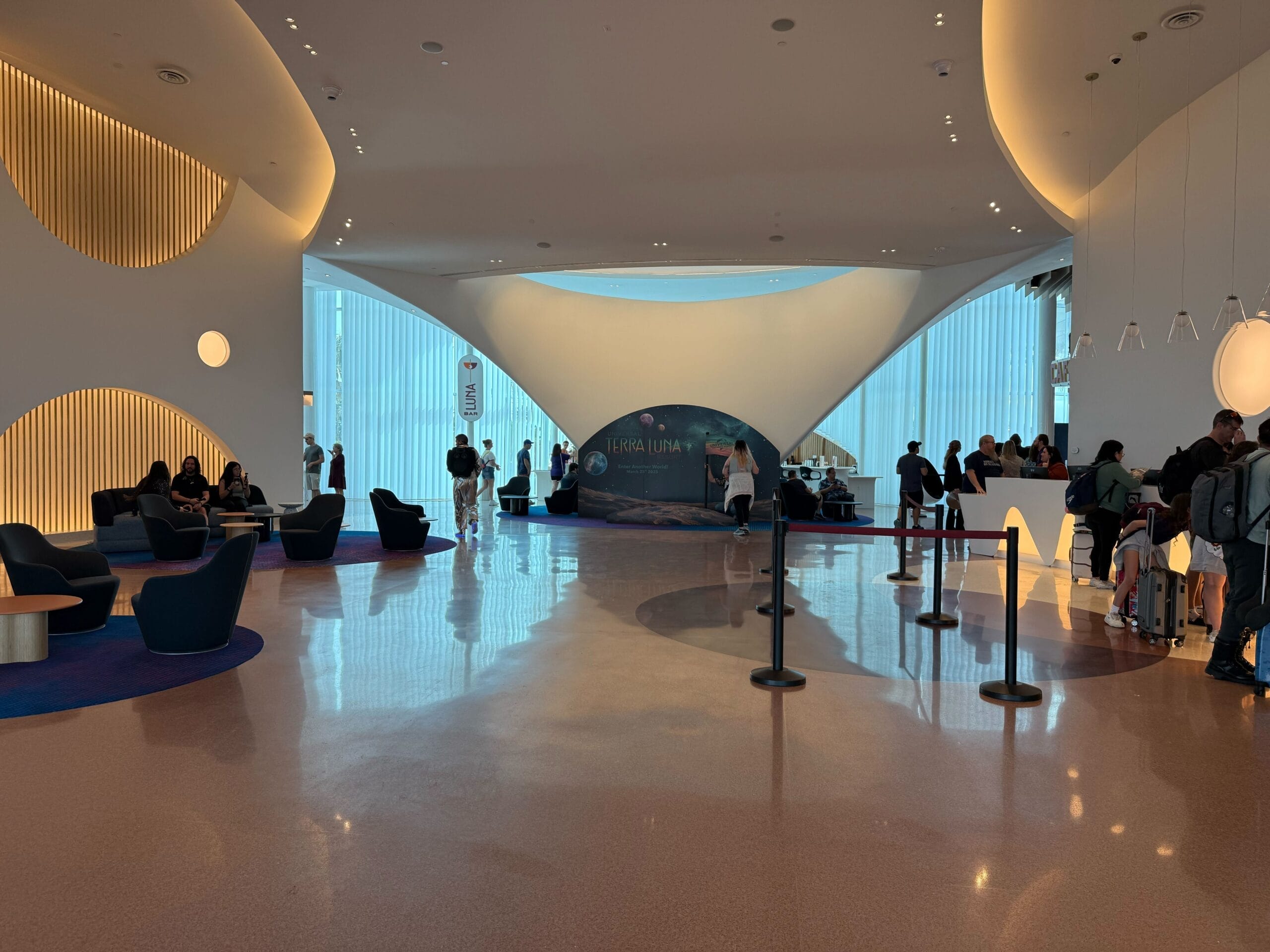 A modern lobby area with curved architecture welcomes guests to the Universal Terra Luna Resort. Seating is on the left, and a reception desk is on the right. A backlit globe display in the background highlights "Lunar," as people wait in line for their adventure to begin.