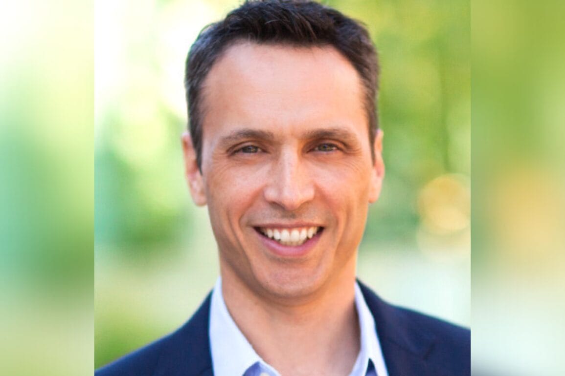 A smiling person in a blazer and shirt stands against a blurred green background, exuding the confident charisma reminiscent of Jimmy Pitaro.