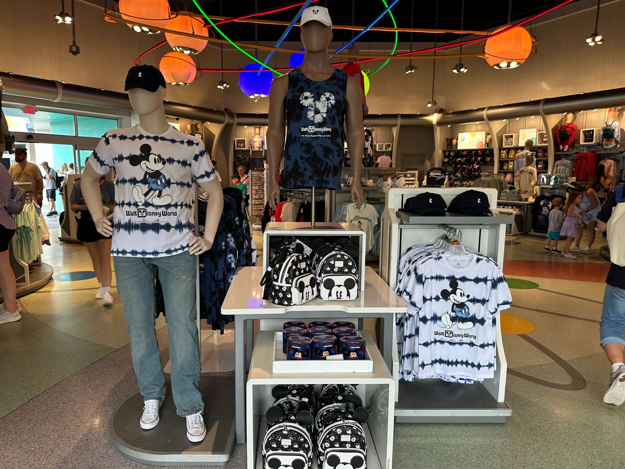 Store display featuring Disney-themed merchandise, including T-shirts, backpacks, and hats, with Mickey Mouse designs. Bright ceiling lights and other shoppers in the background.