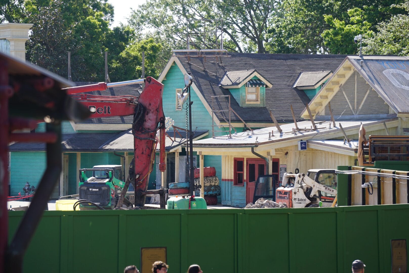 Construction site in front of colorful buildings, with heavy machinery and a green fence in the foreground. Trees are visible in the background.