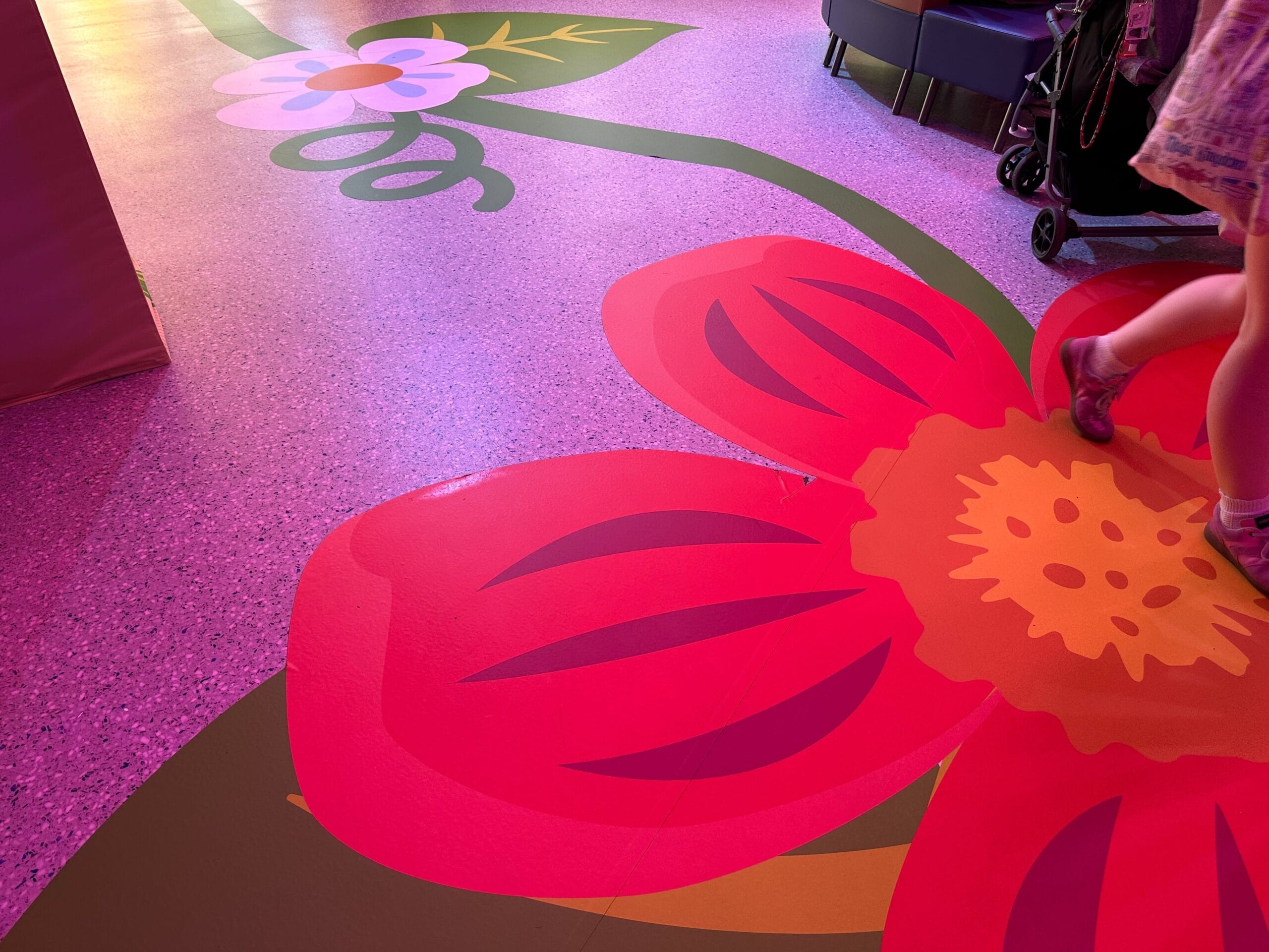 A colorful floor painting features a large red and pink flower with a green stem. A person wearing pink sandals is visible walking by.