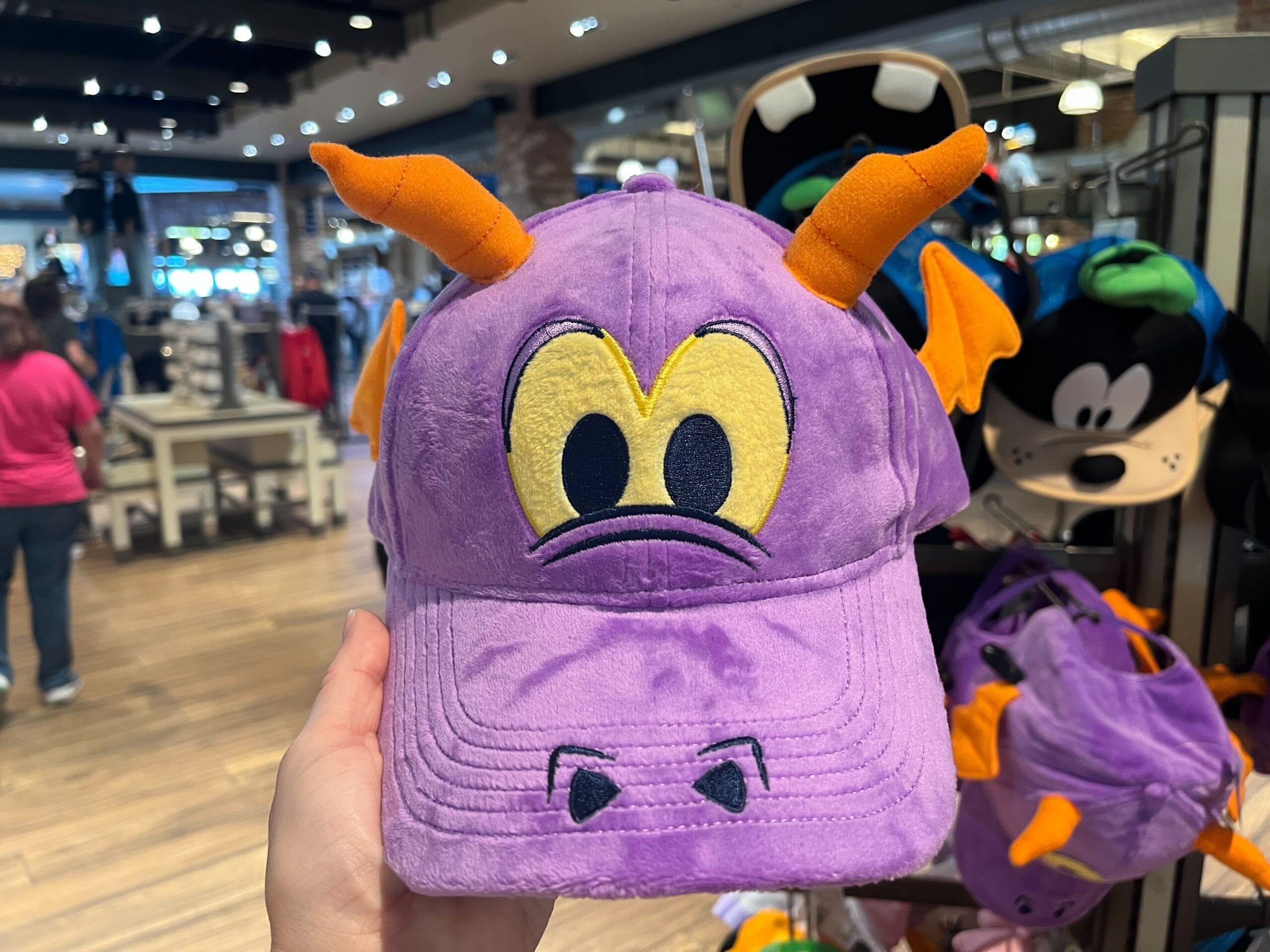 A person holds a purple hat designed to look like a dragon with orange horns and wings in a store.