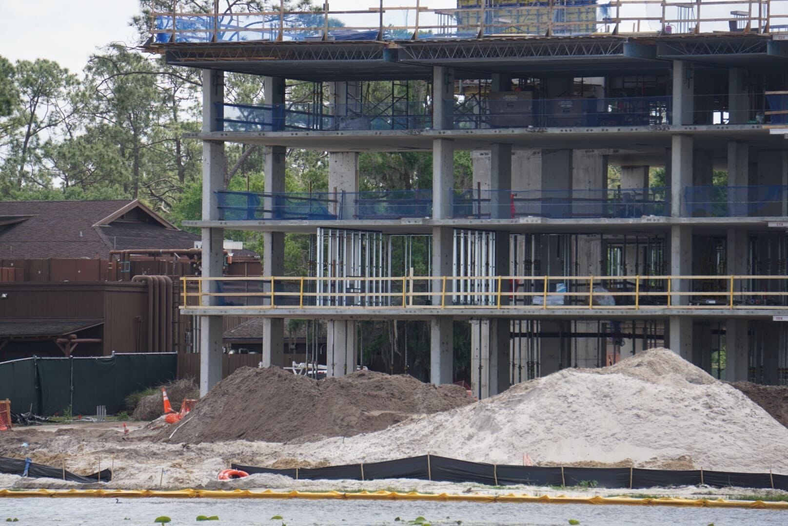 At the construction site, the multi-story building frame rises gracefully, hinting at future developments like the anticipated Disney Lakeshore Lodge. Dirt piles and fencing dominate the foreground, while trees and a wooden structure nestle in the background, completing this dynamic scene.