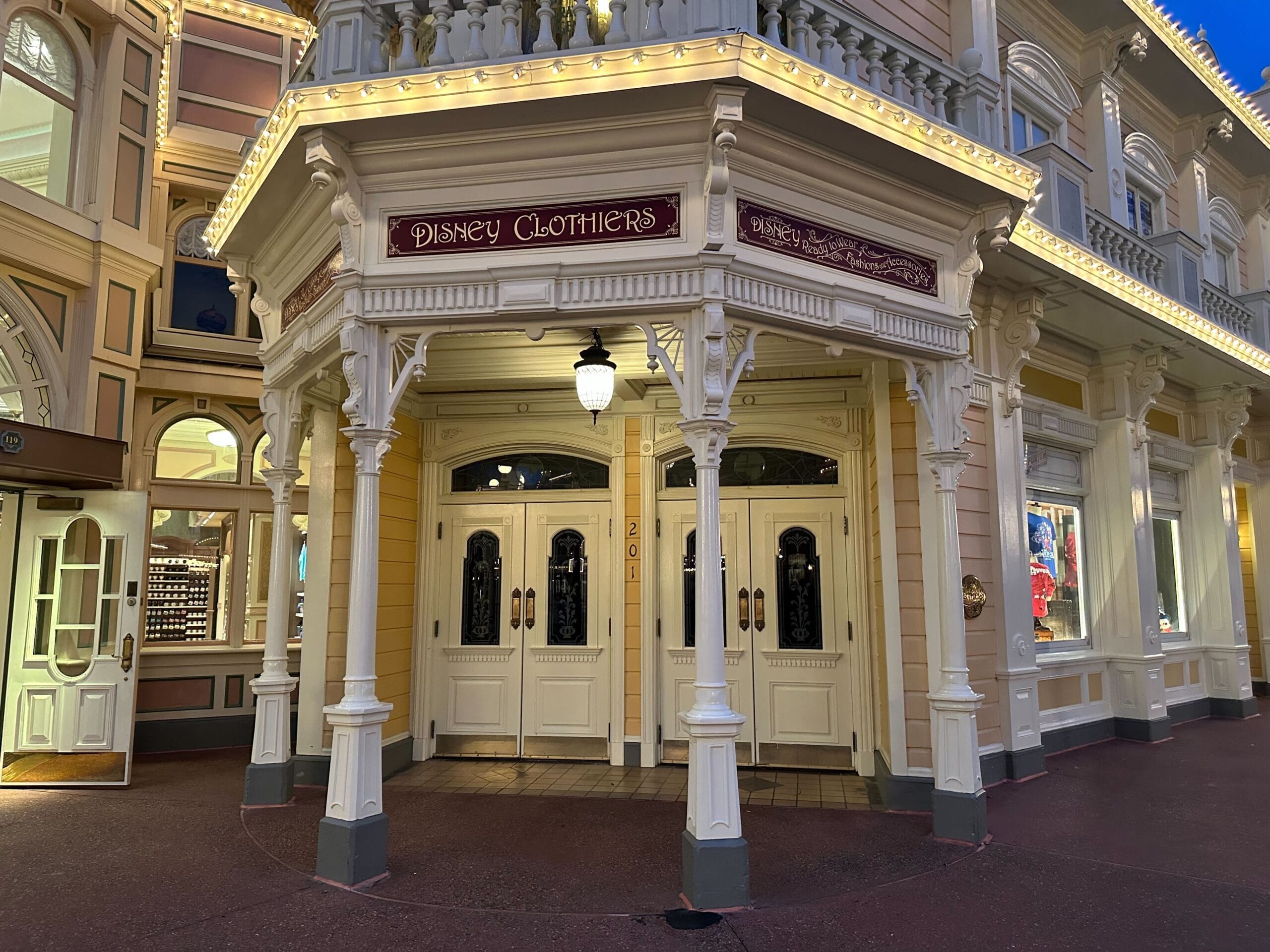 Entrance to Disney Clothiers, a vintage-style building with ornate white columns and trim, warm lighting, and multiple glass doors.
