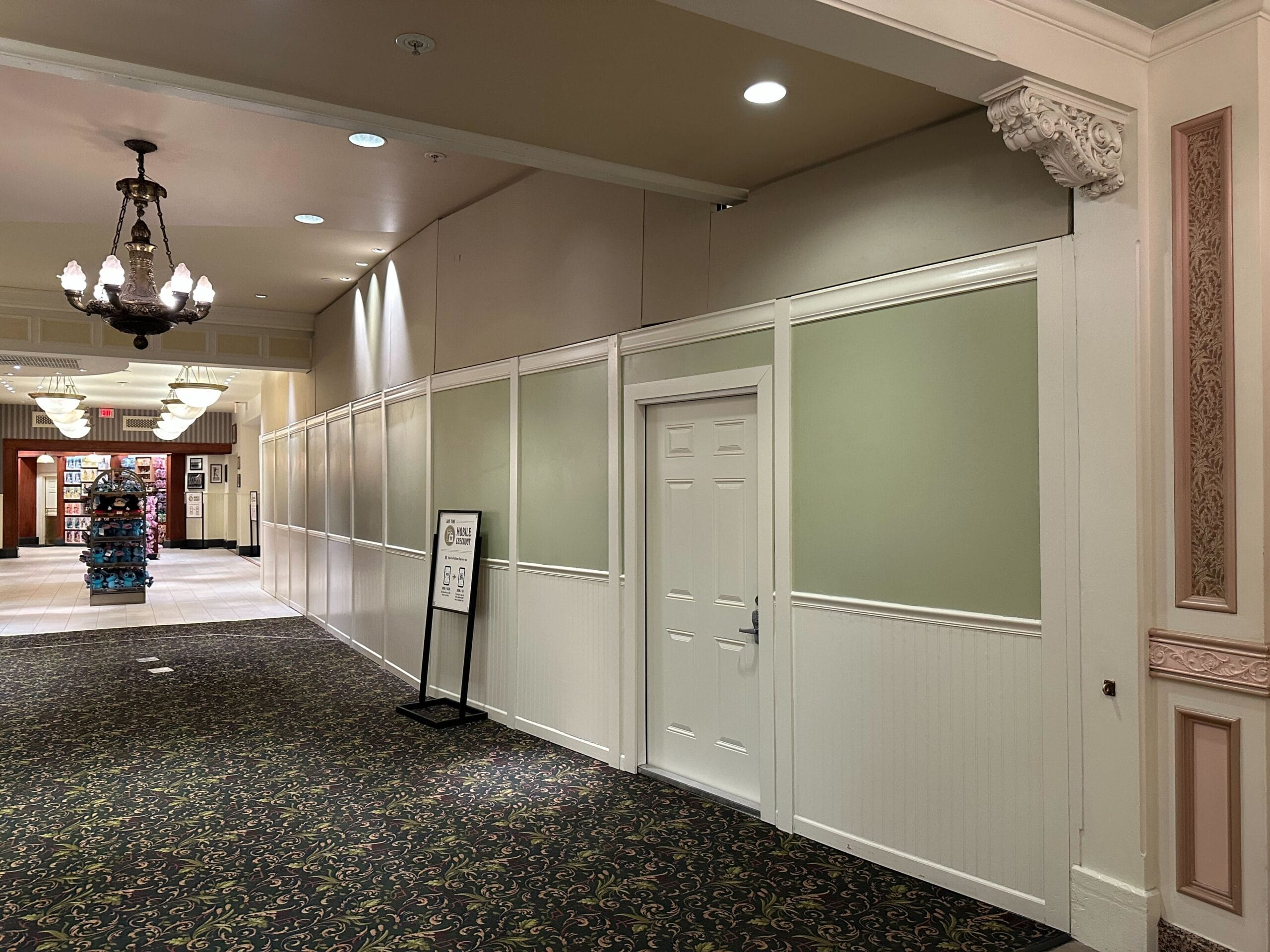A hallway with white-paneled walls and a closed door. A sign stands nearby, with a distant view of a chandelier and a display stand.