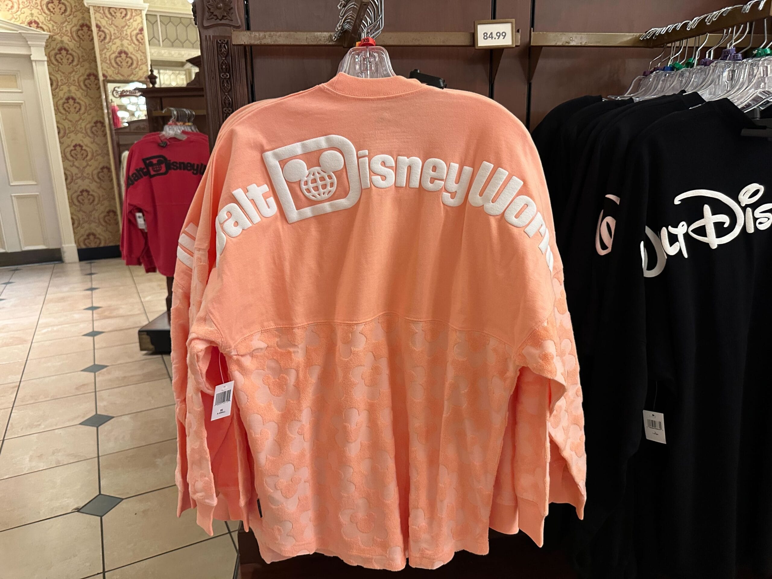 Peach-colored Walt Disney World sweatshirt hanging on a rack, priced at $84.99, with other Disney merch in the background.
