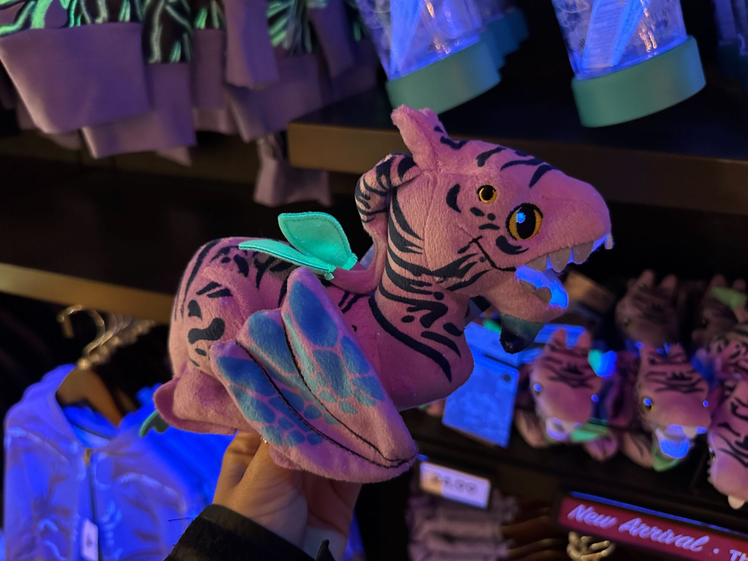 A hand holds a pink dinosaur plush with black stripes, green wings, and light blue accents. Shelves in the background display similar plush toys and clothing.