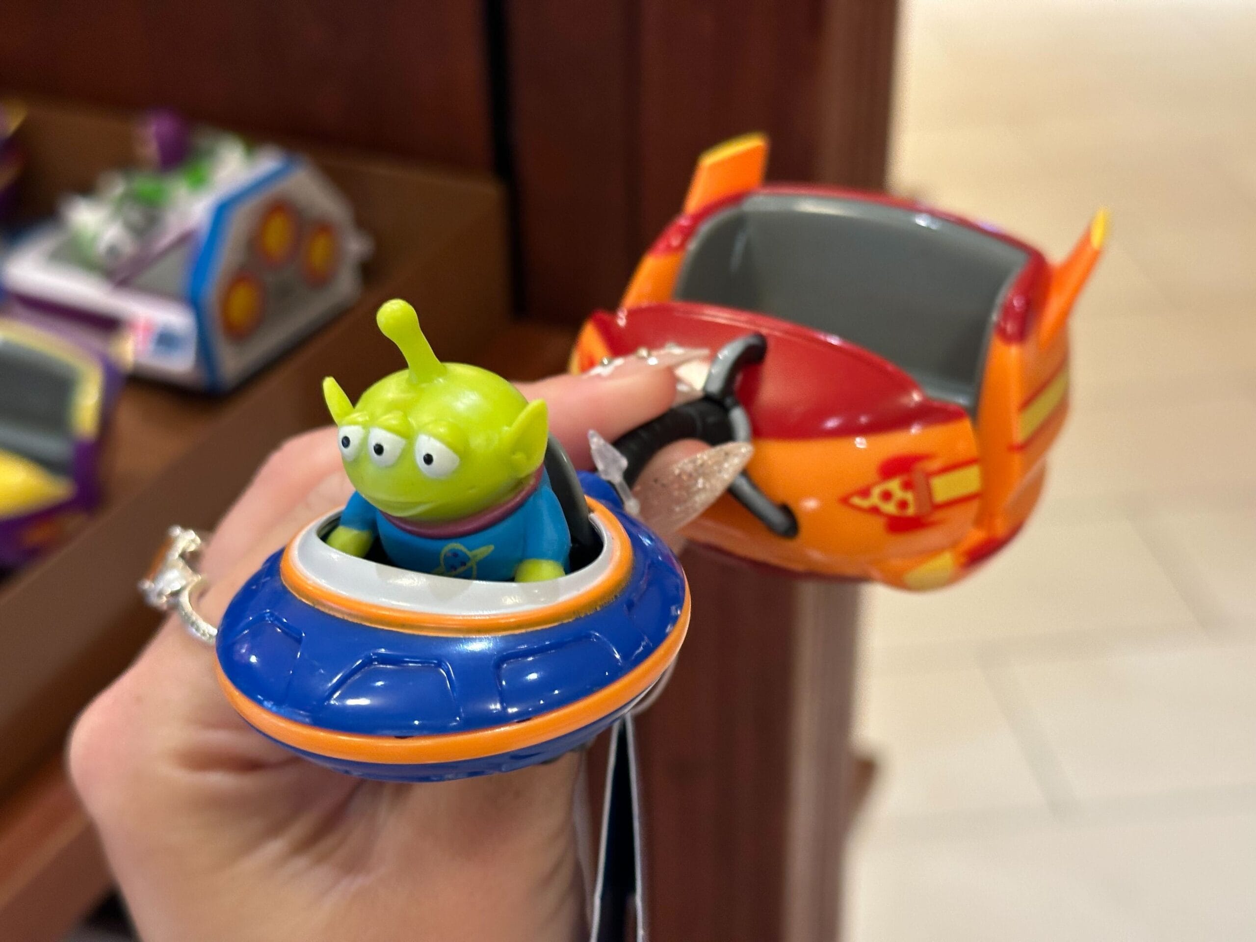 A small green alien toy reminiscent of Walt Disney World's Alien Swirling Saucers sits in a blue and orange UFO-shaped vehicle, held in a person's hand. Another part of this whimsical toy is red and orange with silver wings, capturing the playful spirit of the attraction.