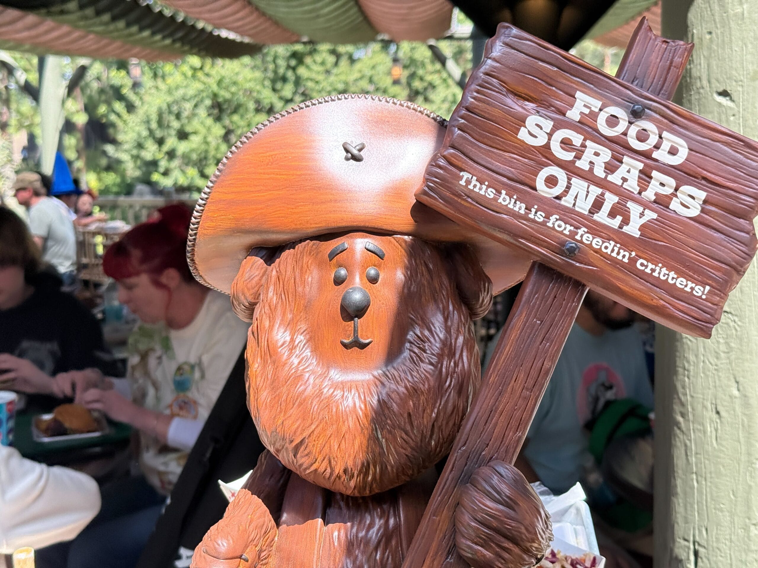 A bear statue from Big Al Carvings, holding a "Food Scraps Only" sign, stands watch as families enjoy meals from Hungry Bear Barbecue in the lively atmosphere of Disneyland Park.