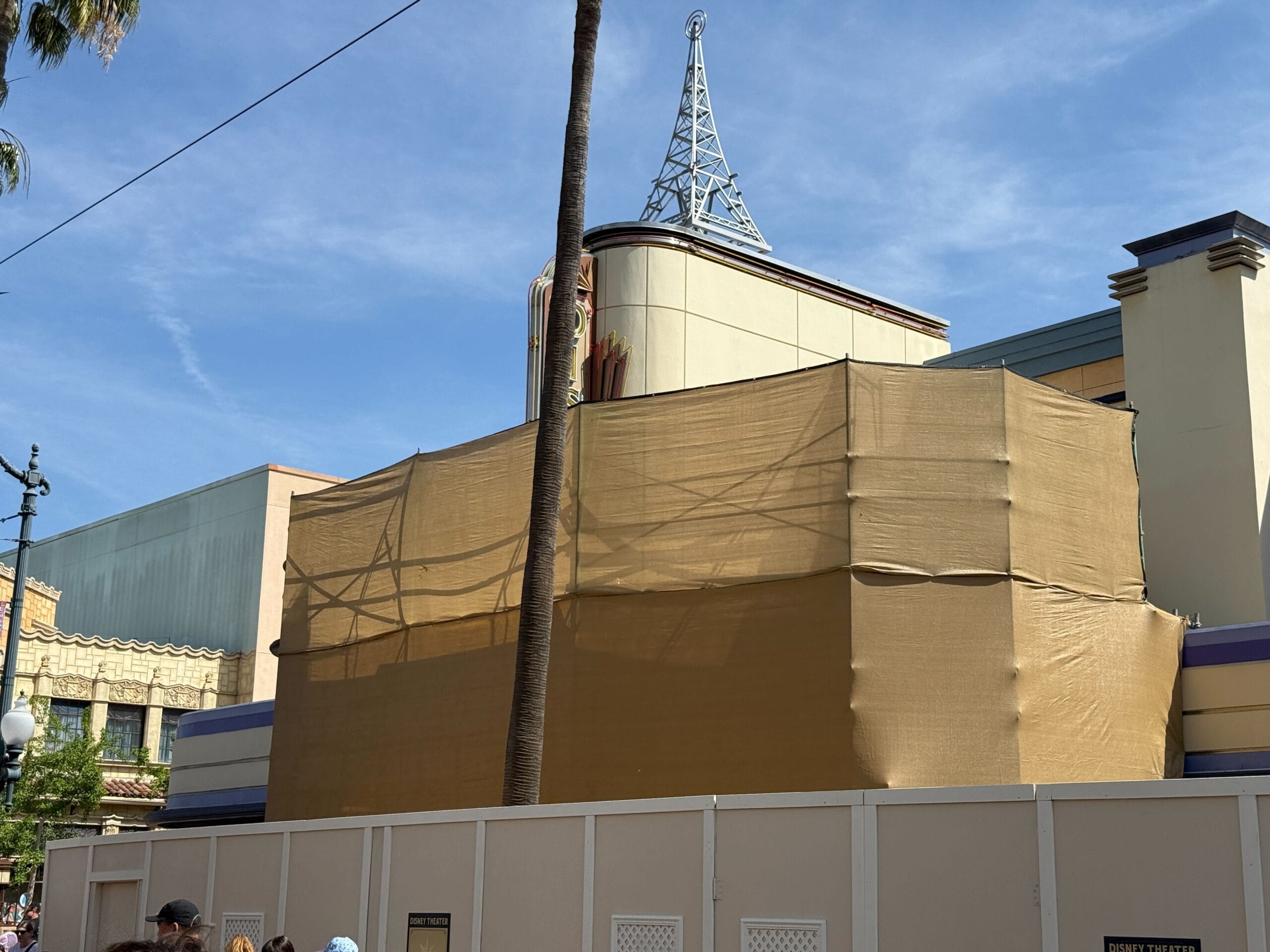 The building under construction, draped in beige tarp with a decorative spire on top, hints at the magic behind Disneyland's latest transformation. A palm tree sways against the blue sky, while construction walls keep the mystery of Disney's new show concealed until the grand reveal.