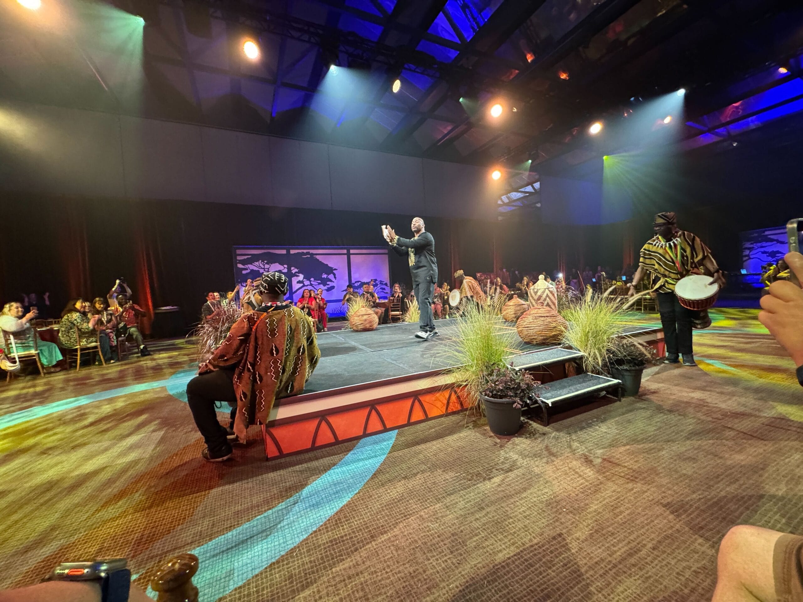 A performer takes center stage with dancers and musicians in traditional attire during a lively Feast of the Lion King event. Bright stage lights illuminate the scene as the audience, eagerly seated around, is transported to the heart of the Pride Lands.