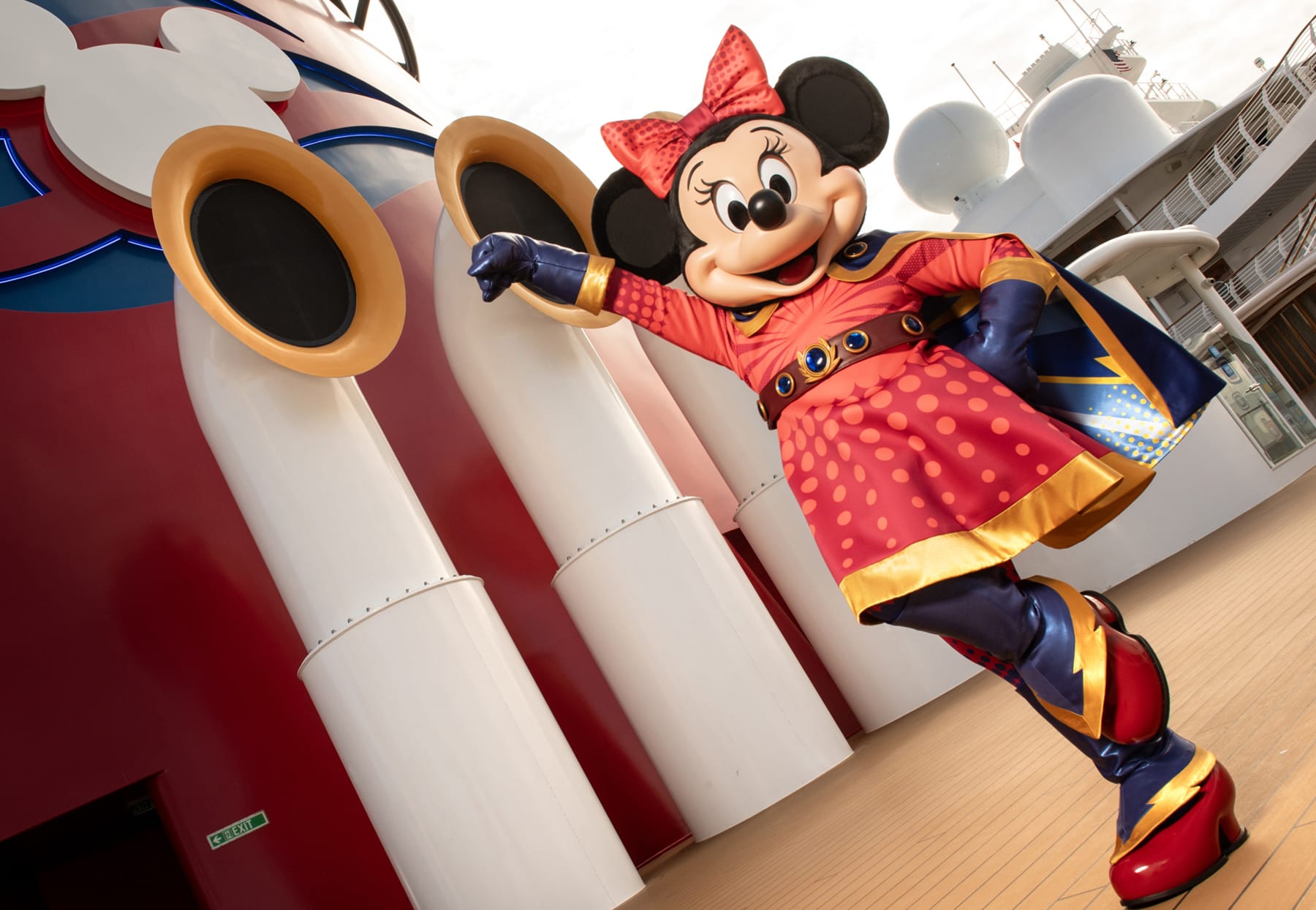 A person in a Minnie Mouse superhero costume poses energetically on a ship deck, next to large red and white smokestacks.