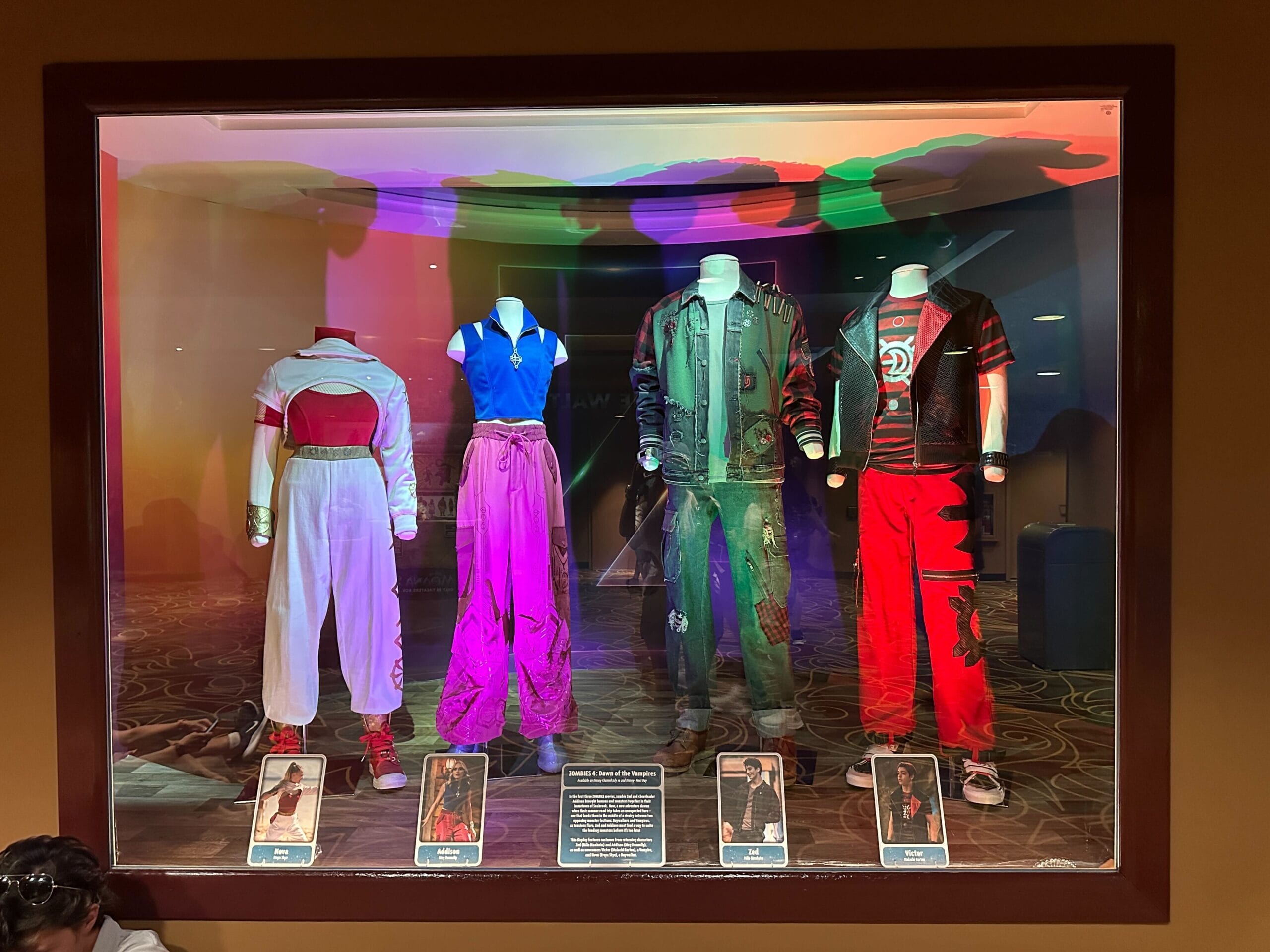 Four colorful costumes from a film are displayed in a glass case with character cards and descriptions below each outfit.