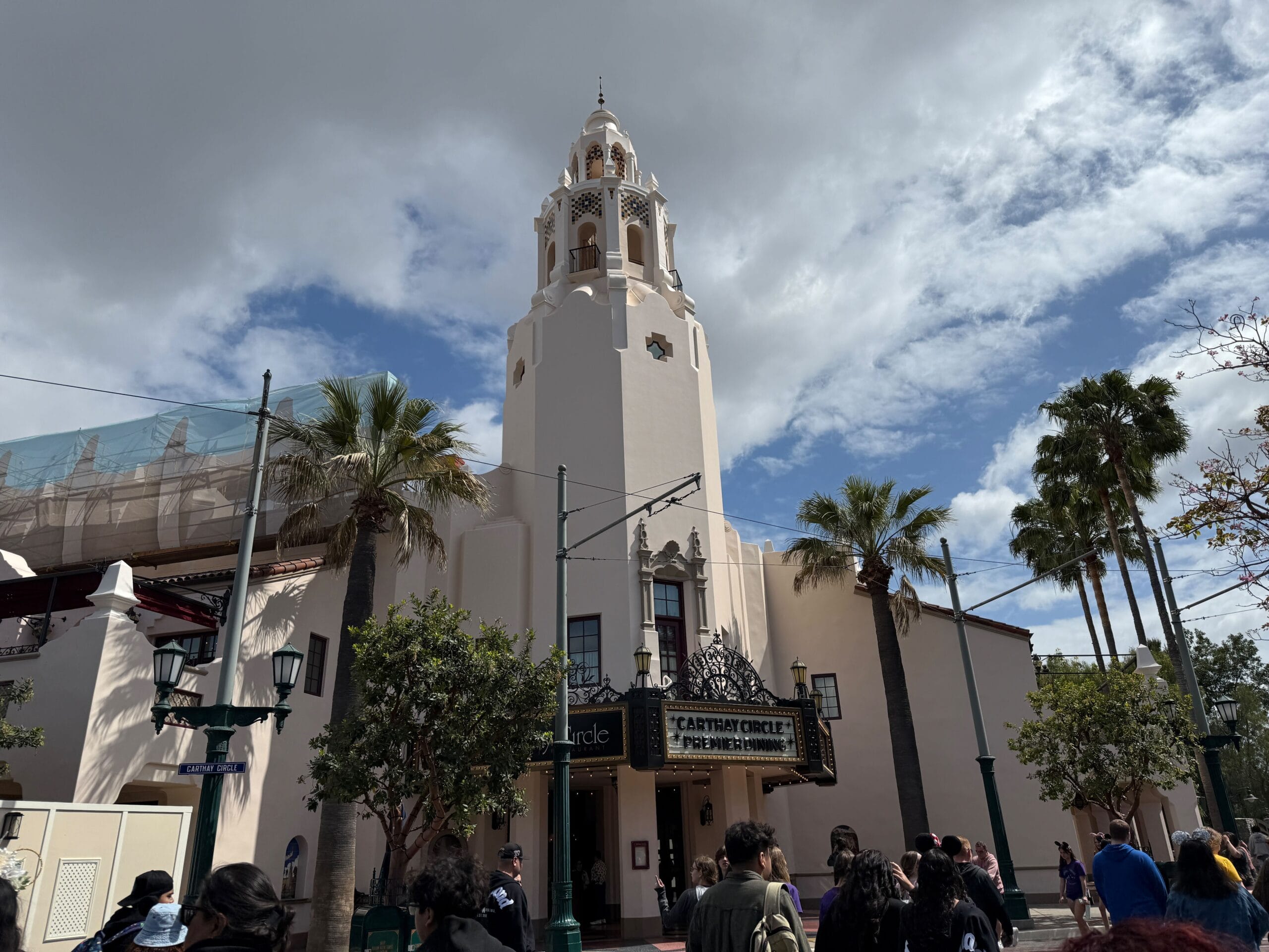 A tall white building with a tower, palm trees, and people strolling by Disney’s Carthay Circle Restaurant.
