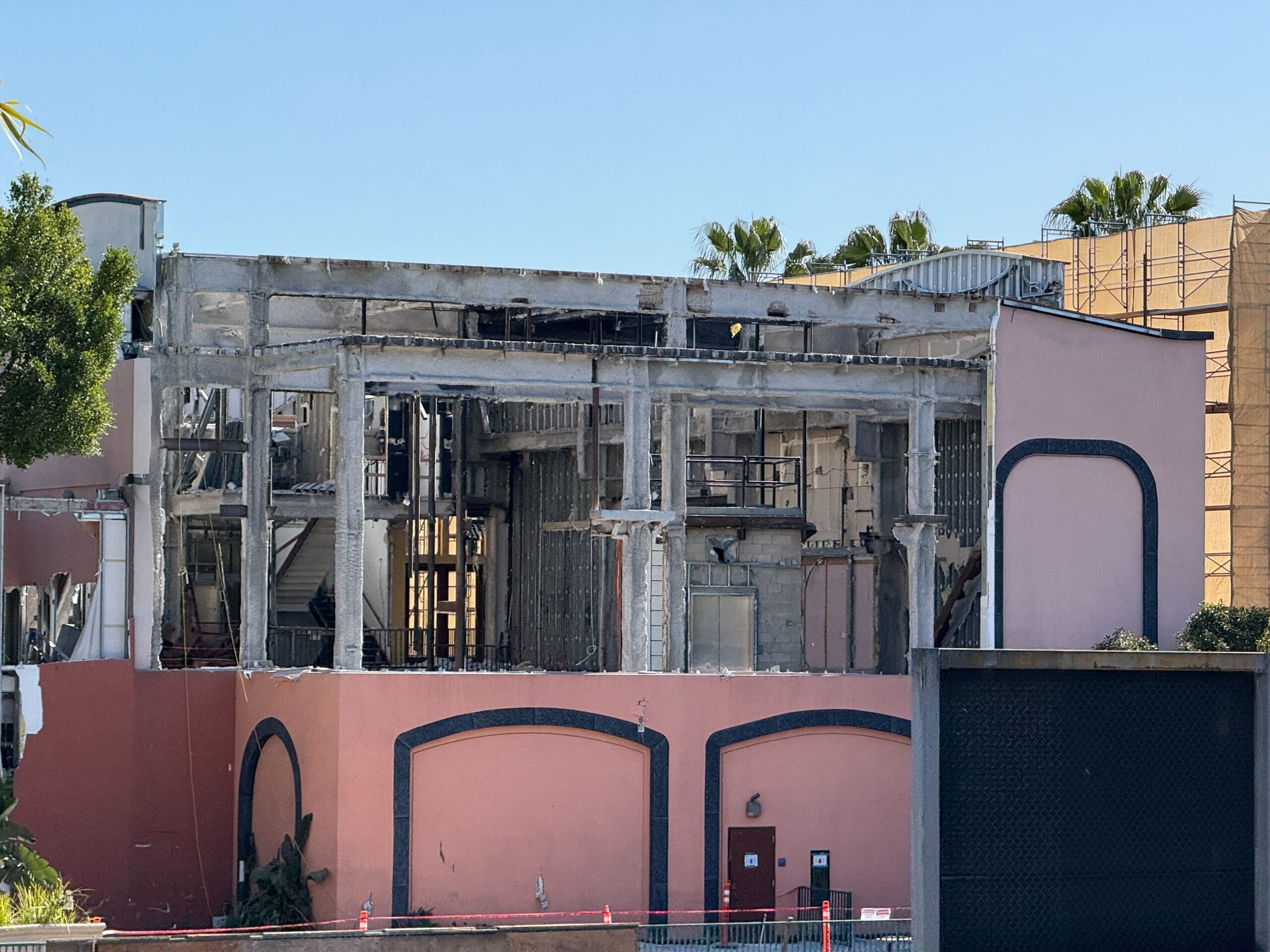 Partially demolished building with exposed steel beams and interior. The pink exterior facade remains intact. Surrounding trees and buildings are visible under a clear blue sky.