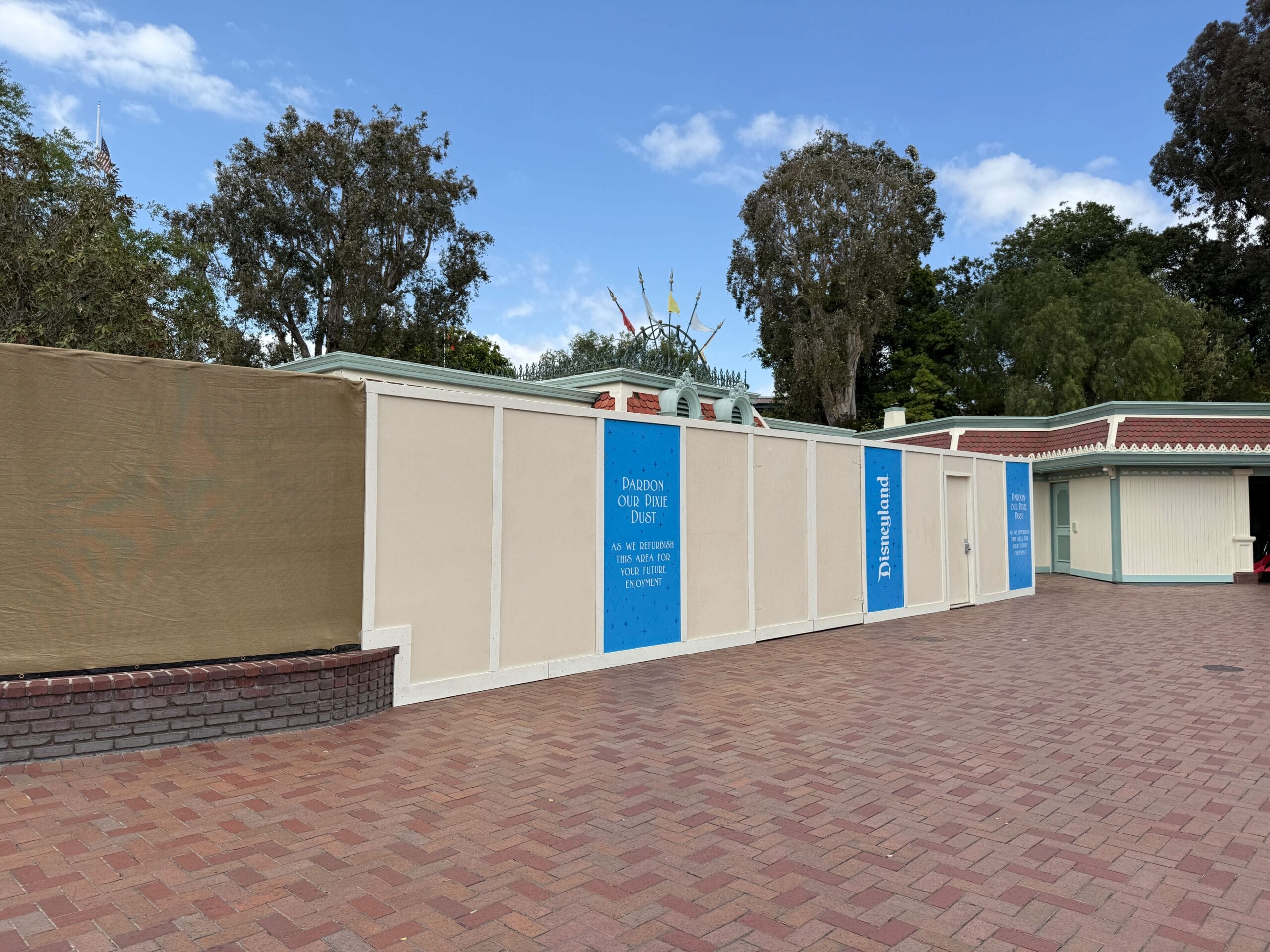 Construction walls with blue Disneyland entrance signs block a brick walkway, with trees and a building in the background.