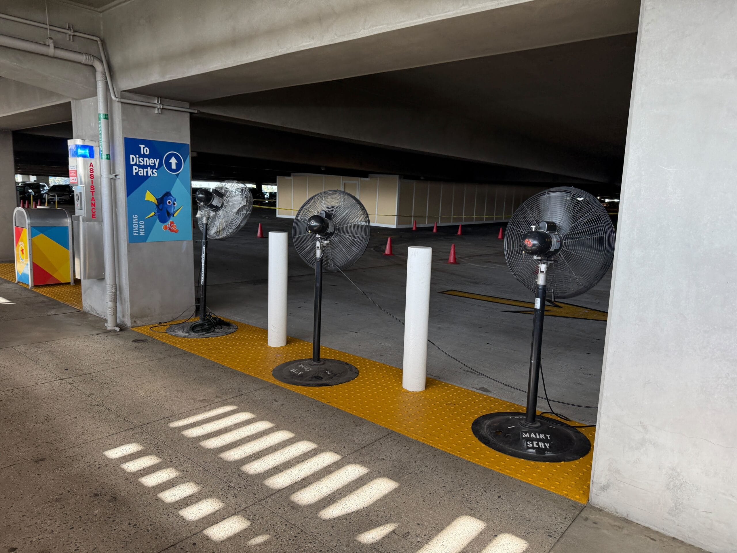 Three large fans are positioned in a row near the entrance of a parking garage. A directional sign points "To Disney Parks.