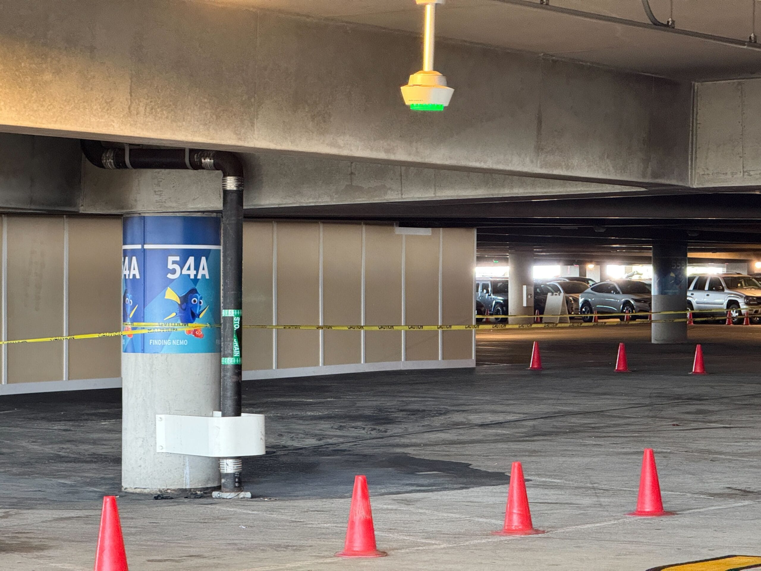 Indoor parking garage area with red cones blocking sections, overhead sensor with green light, visible numbered column 54A, and partially occupied parking spaces in the background.