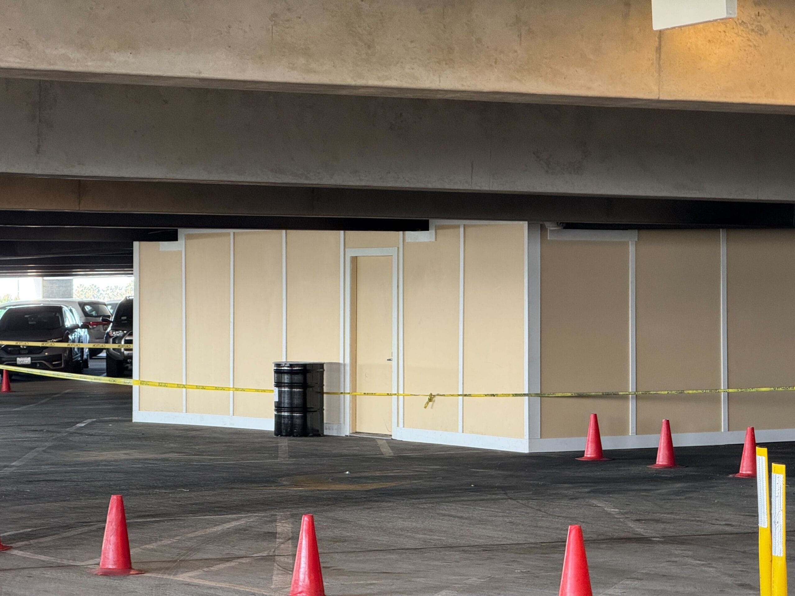 A construction area in a parking garage is marked by caution tape and traffic cones. A temporary wall encloses the space with a visible door.