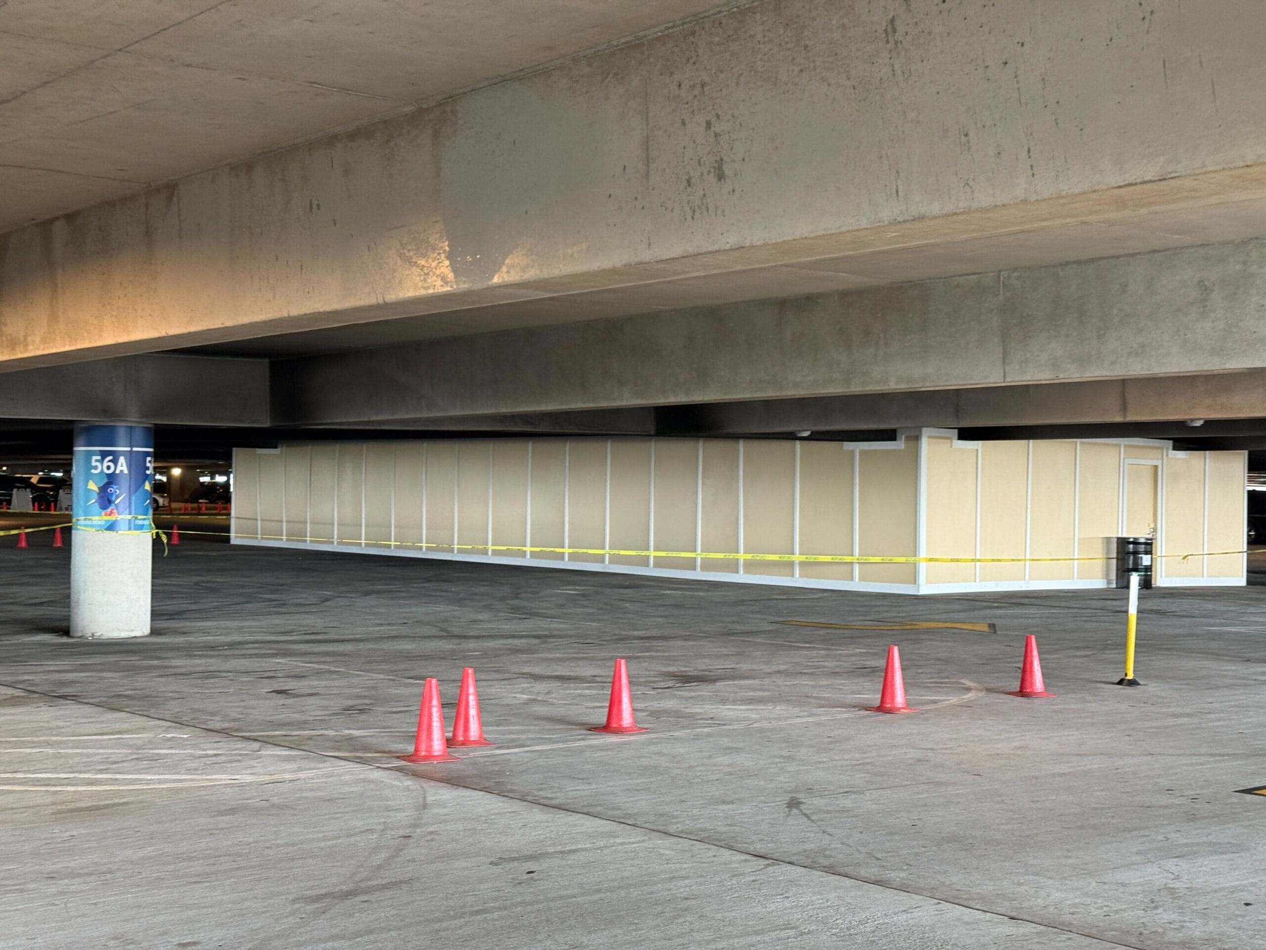 A construction area under a concrete parking structure is enclosed by beige panels, marked off with yellow tape and surrounded by orange traffic cones. A column is labeled "56A.