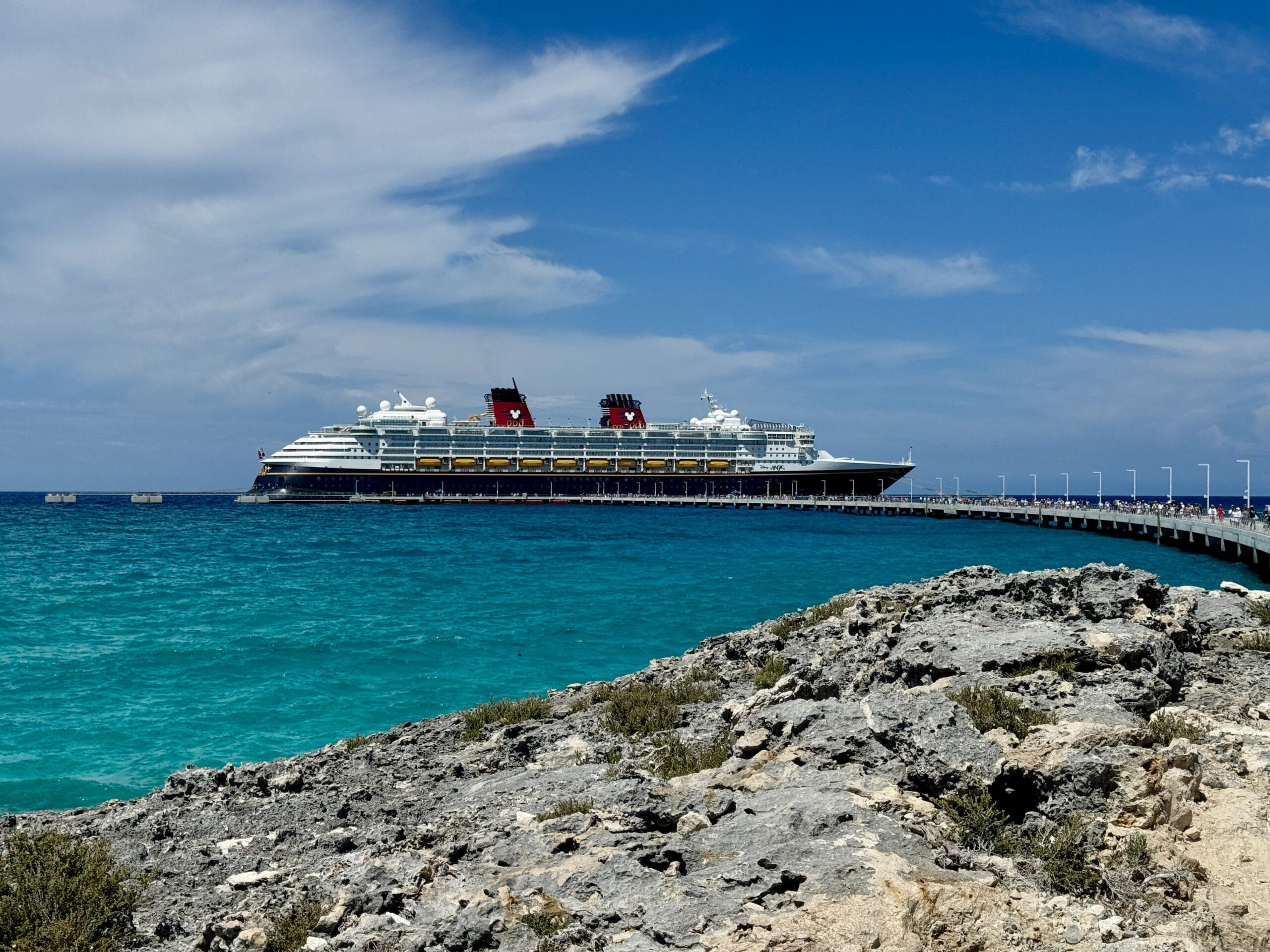 A large Disney Magic cruise ship rests beside the pier on a clear day, with turquoise waters in the foreground and a partly cloudy sky above.